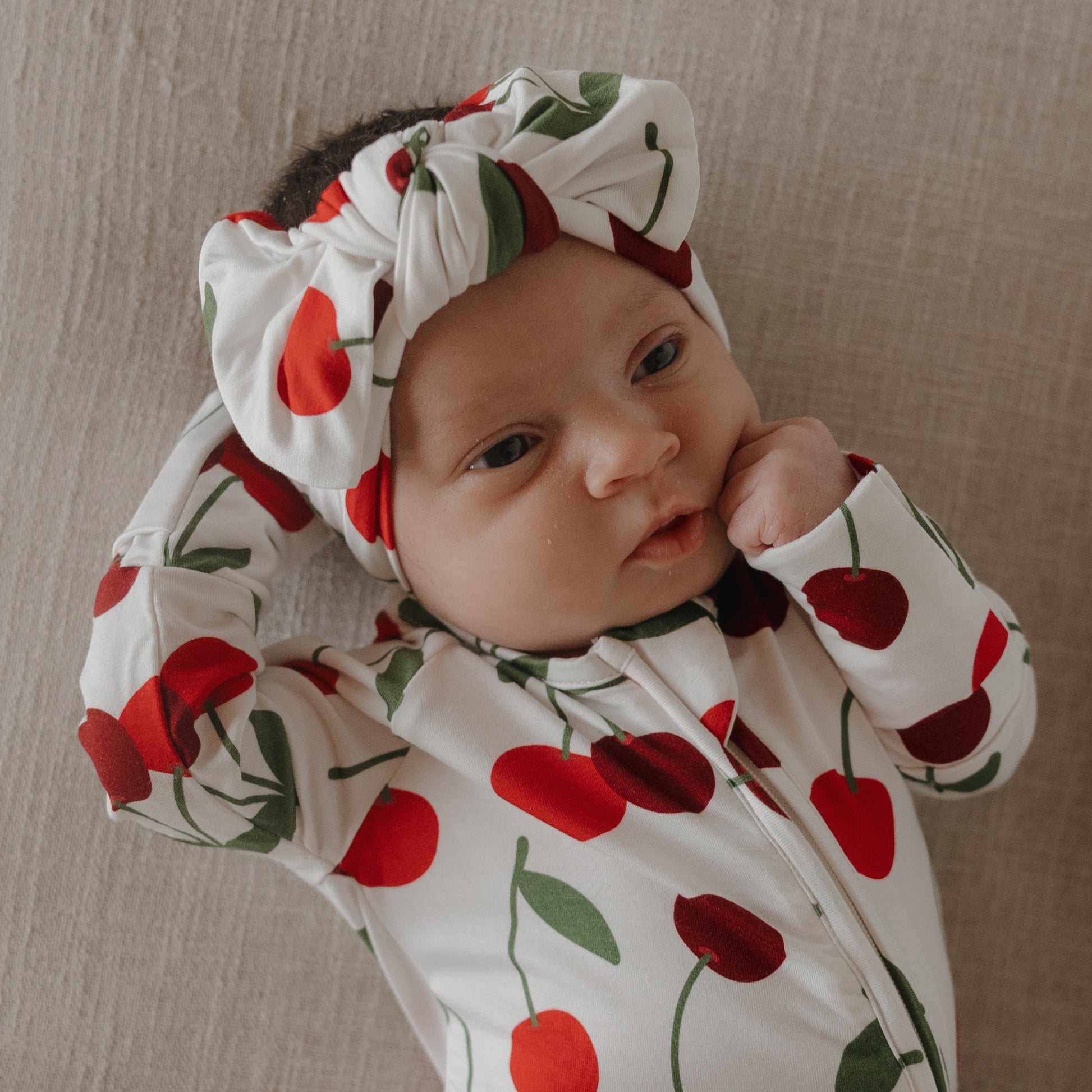 A baby in a white outfit wears the forever french baby Bamboo Head Wrap | Cherry, featuring a red cherry pattern, while lying on beige fabric with one hand resting on their head and the other near their cheek.
