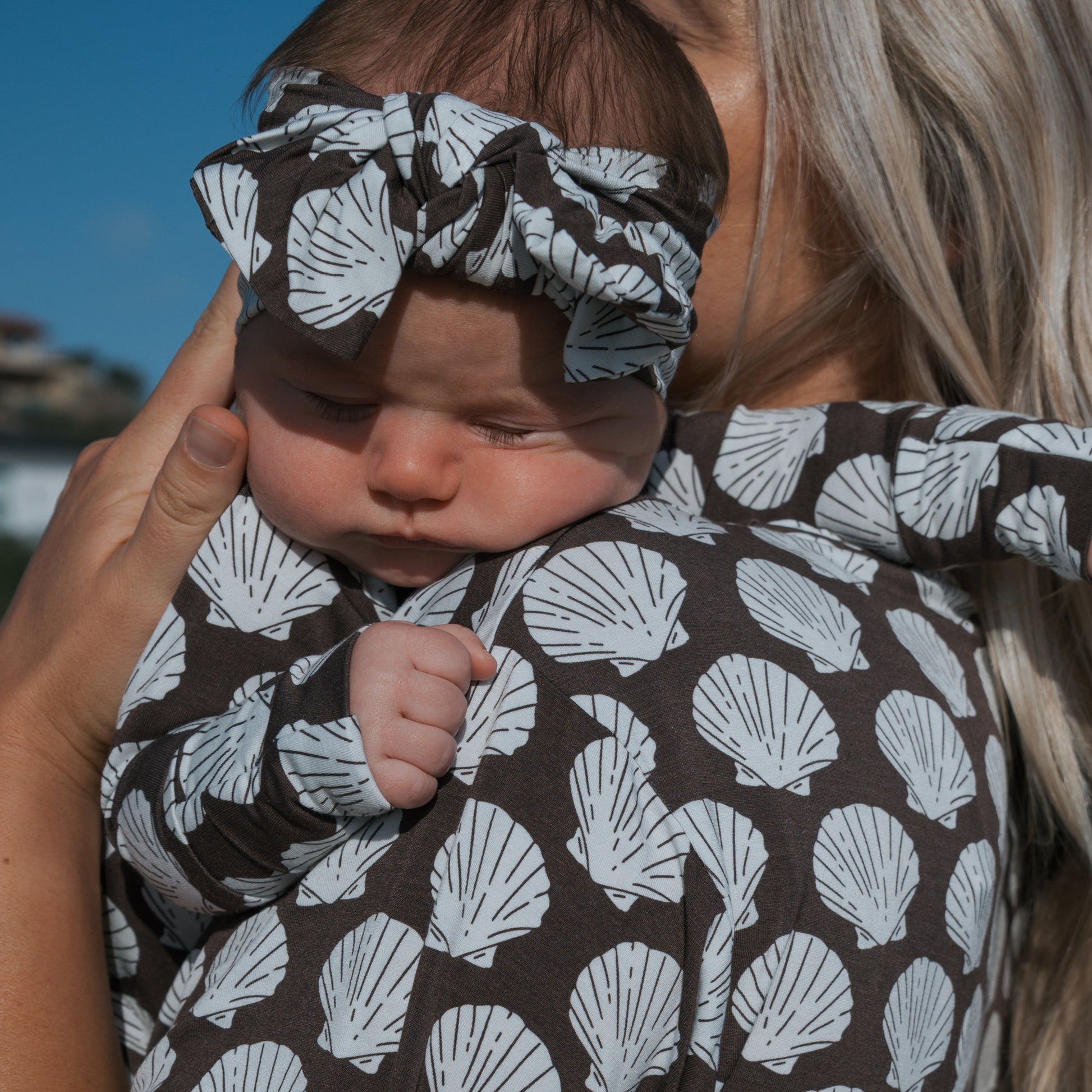 A baby in a matching brown forever french baby outfit and a Bamboo Head Wrap | Seashells rests on an adult’s shoulder, with the adult's long blonde hair and gentle hand visible.
