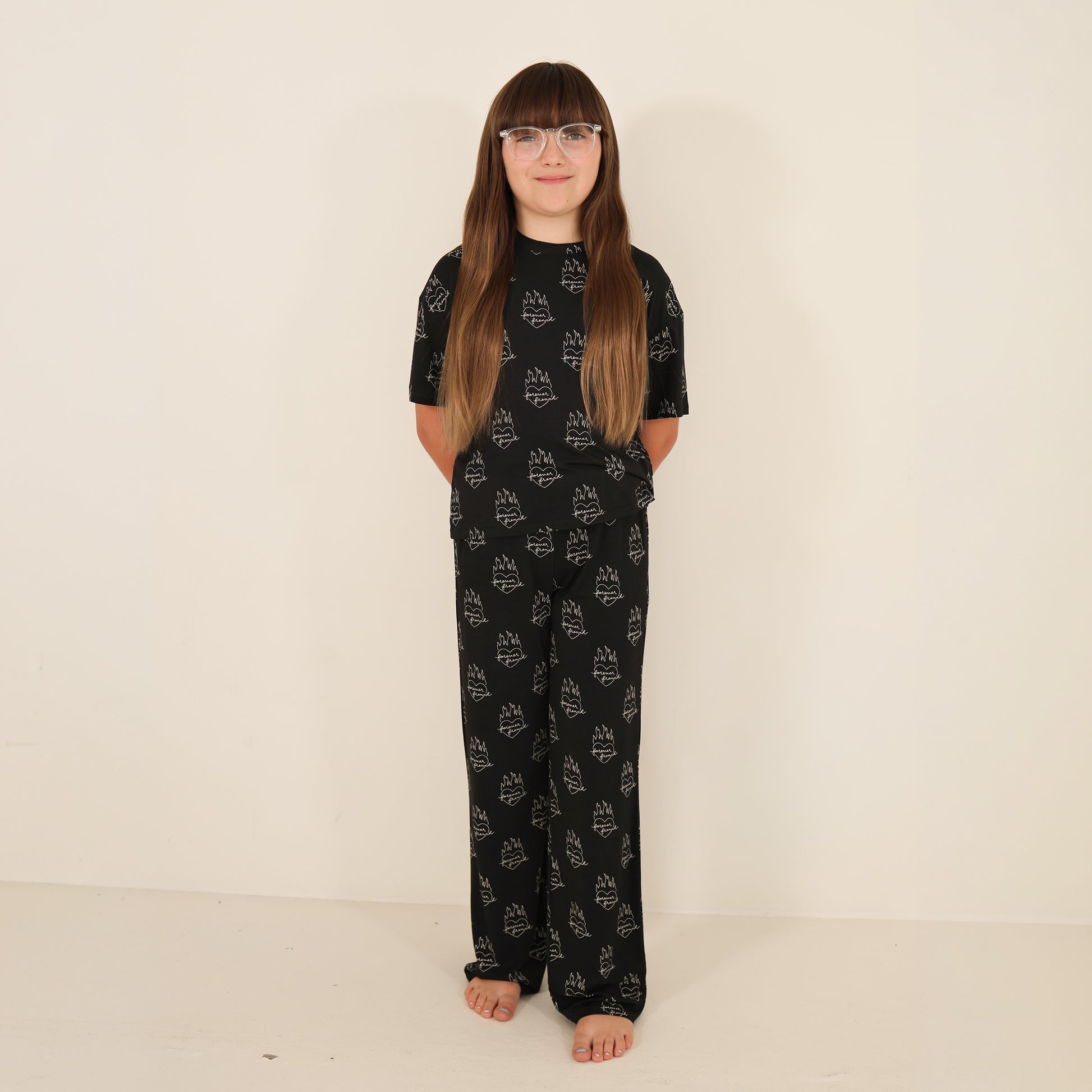 A child with long brown hair and glasses stands barefoot against a white wall, smiling in the forever french baby Child Boxy Tee Lounge Set | Flaming Hearts—breathable, comfortable kids sleepwear with a playful pattern.