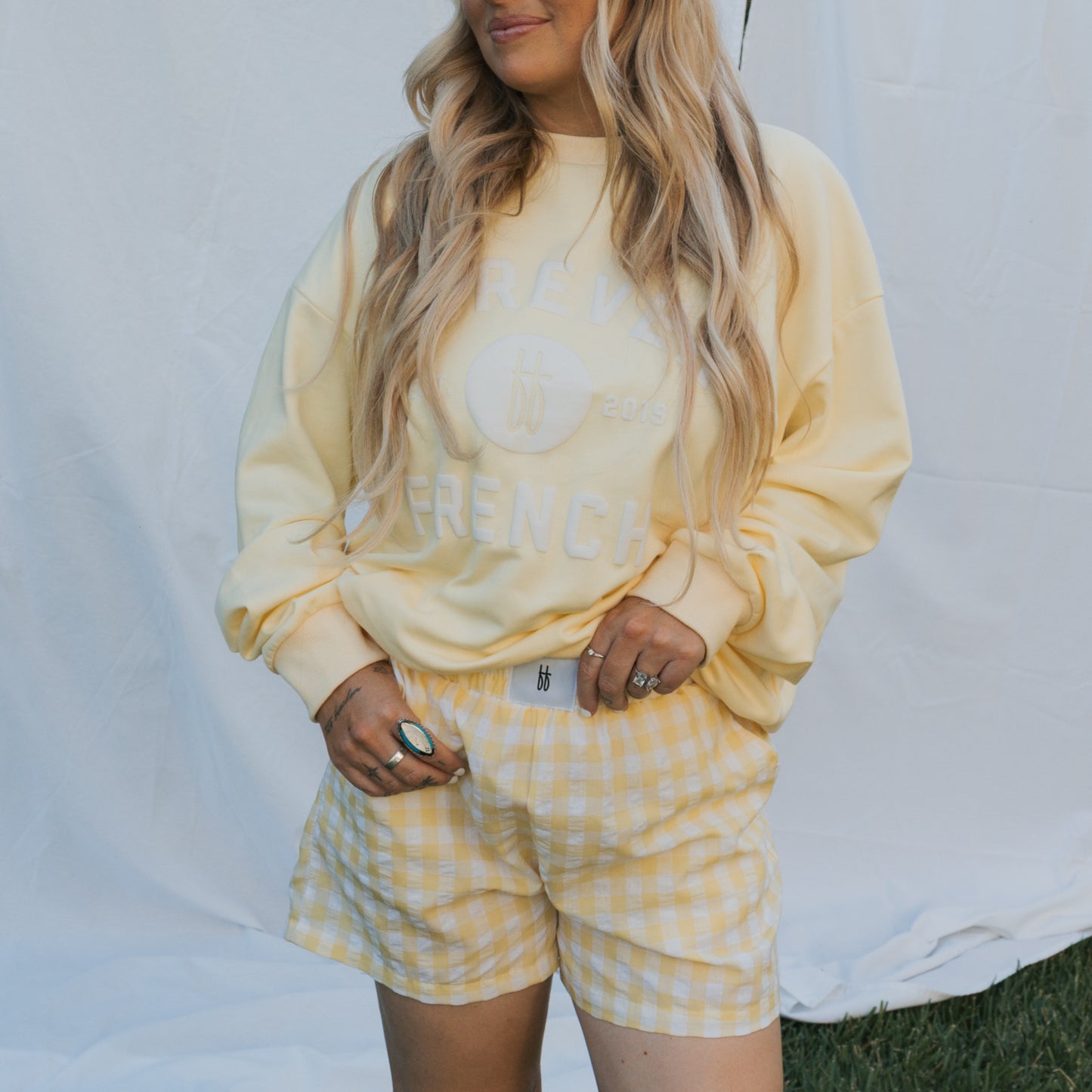 A woman with long wavy blonde hair models forever french baby's Women's Boxer Shorts in Citron, posing with hands in her pockets before a white fabric backdrop on a grassy lawn.