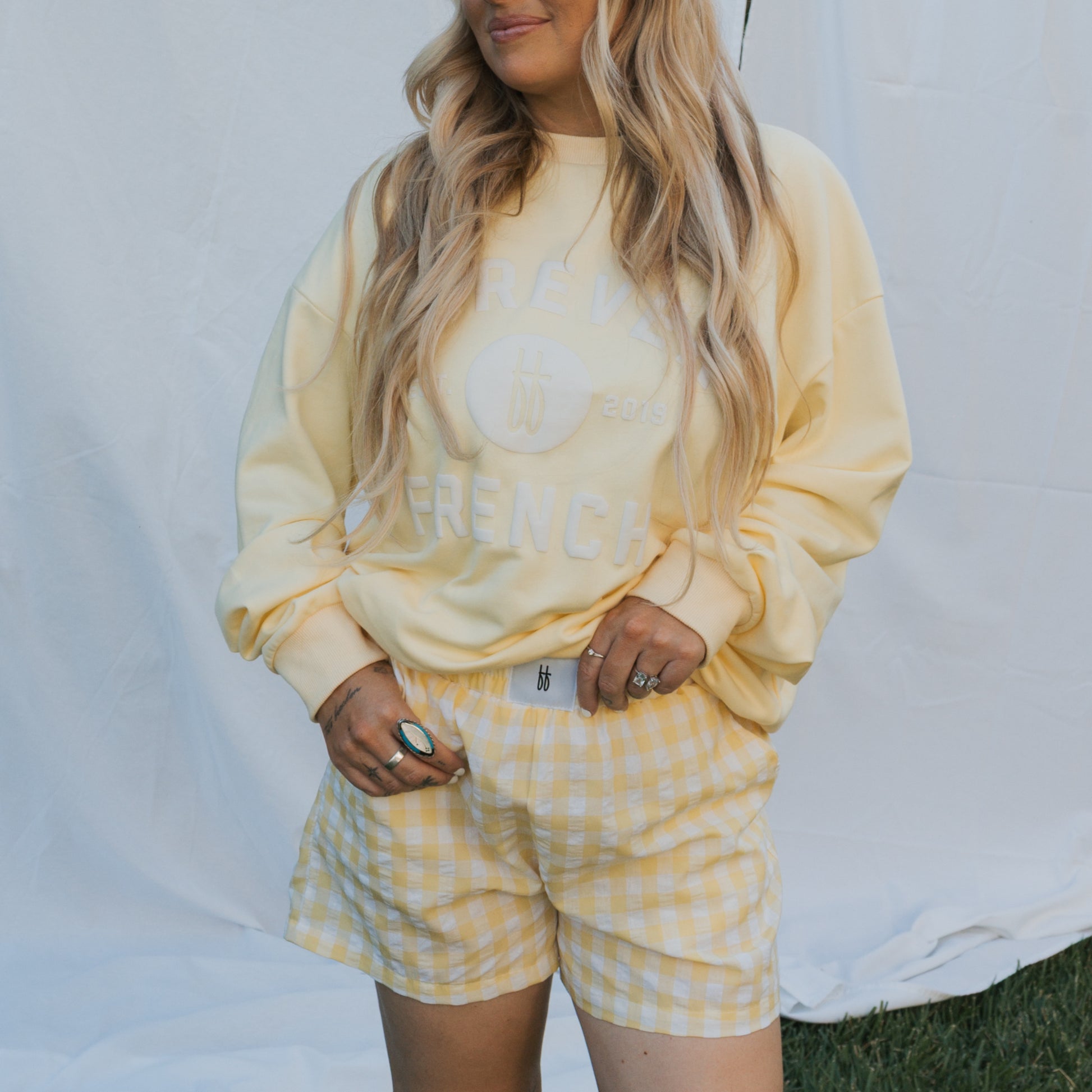 A woman with long wavy blonde hair models forever french baby's Women's Boxer Shorts in Citron, posing with hands in her pockets before a white fabric backdrop on a grassy lawn.