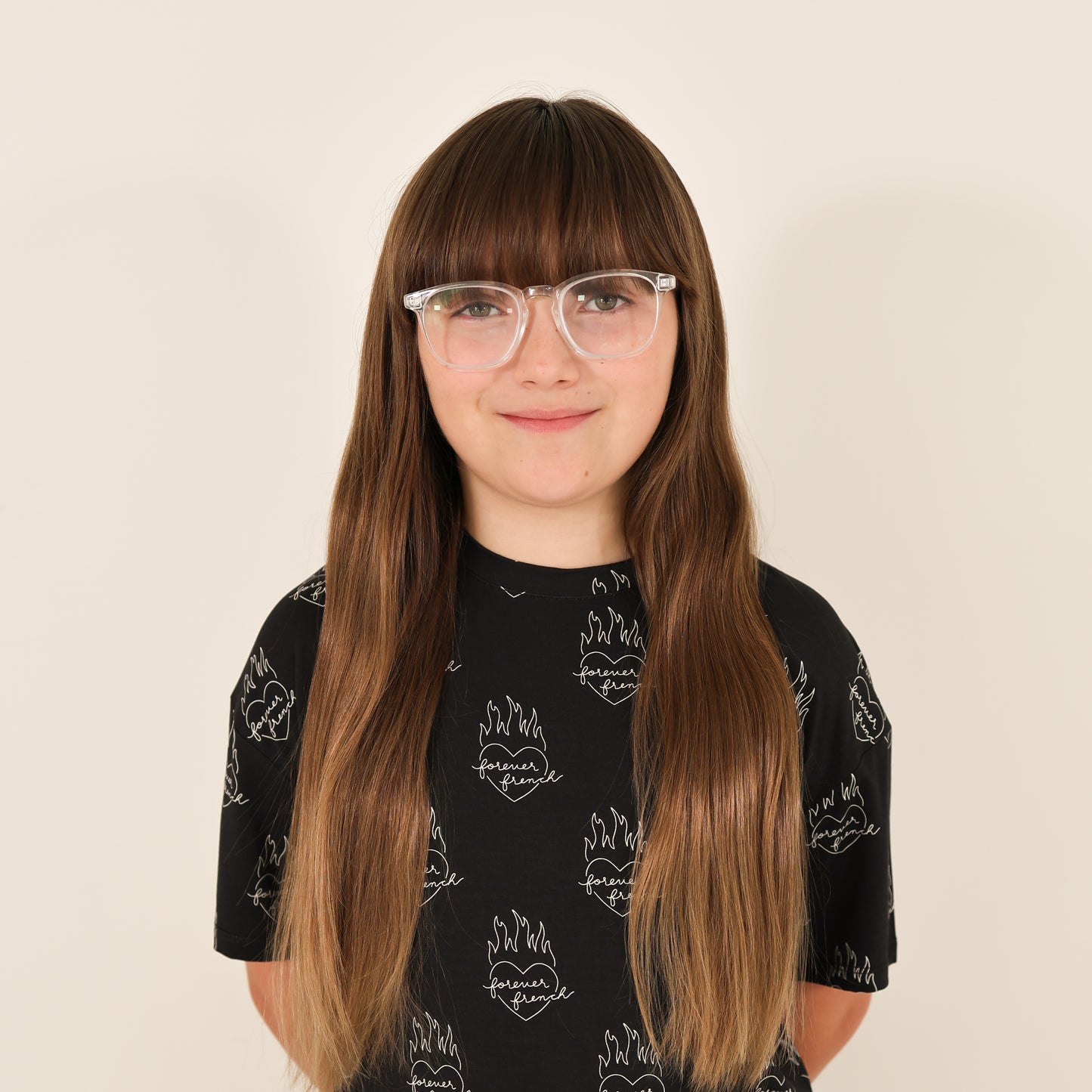 A young person with long brown hair, bangs, and clear glasses wears the forever french baby Child Boxy Tee Lounge Set | Flaming Hearts—a black shirt with a white flame and text design—standing against a plain light background, smiling slightly.