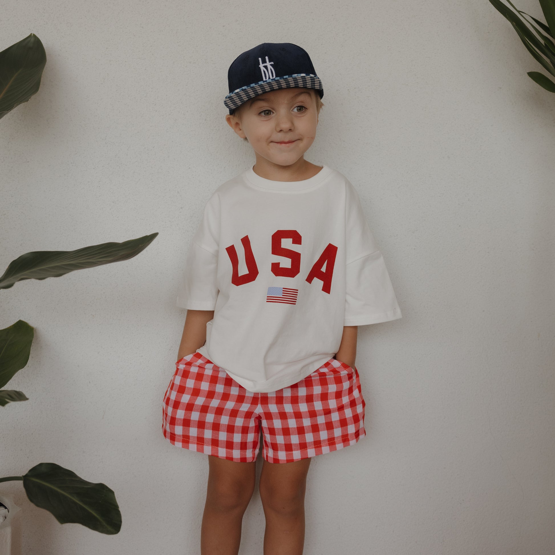 A young child in a navy blue cap, white "USA" T-shirt, and forever french baby Child Boxer Shorts | Scarlet Gingham, stands with hands in pockets against a plain wall flanked by green plants.
