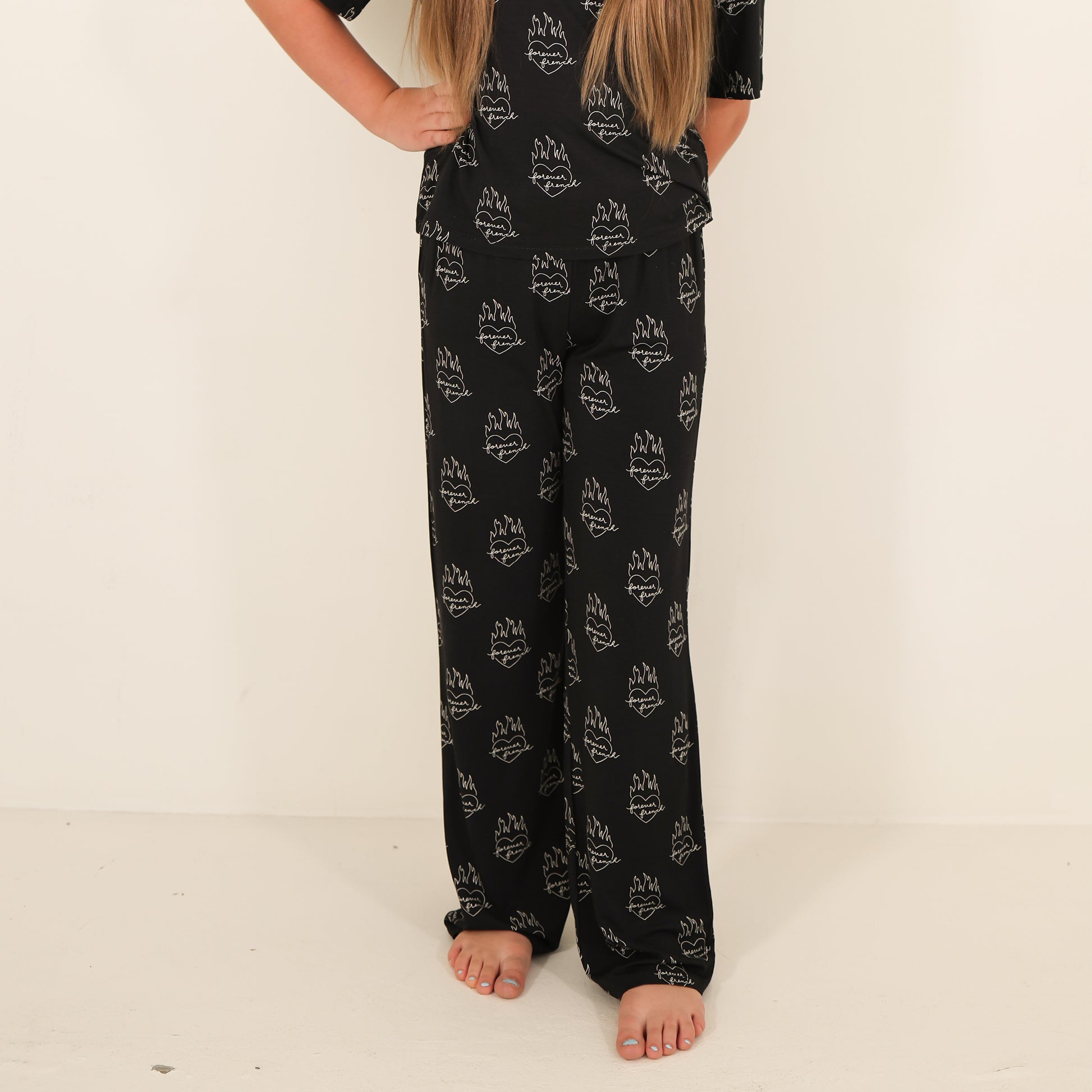 A person with long hair is wearing the forever french baby Child Boxy Tee Lounge Set in Flaming Hearts, standing barefoot on a light-colored floor against a plain wall. Only part of their upper body is visible.
