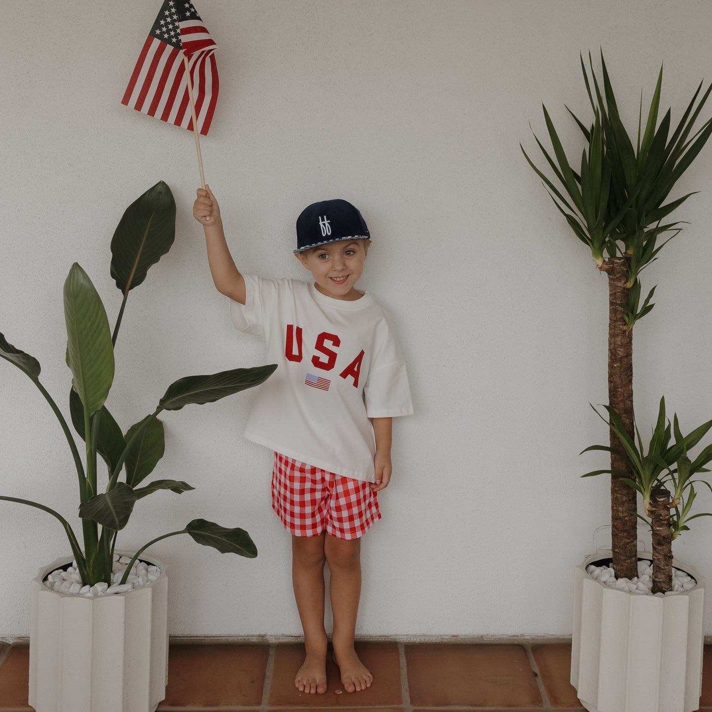 A young child stands barefoot between two potted plants, smiling against a white wall, wearing a "USA" shirt, blue cap, and forever french baby's Child Boxer Shorts | Scarlet Gingham while holding an American flag.