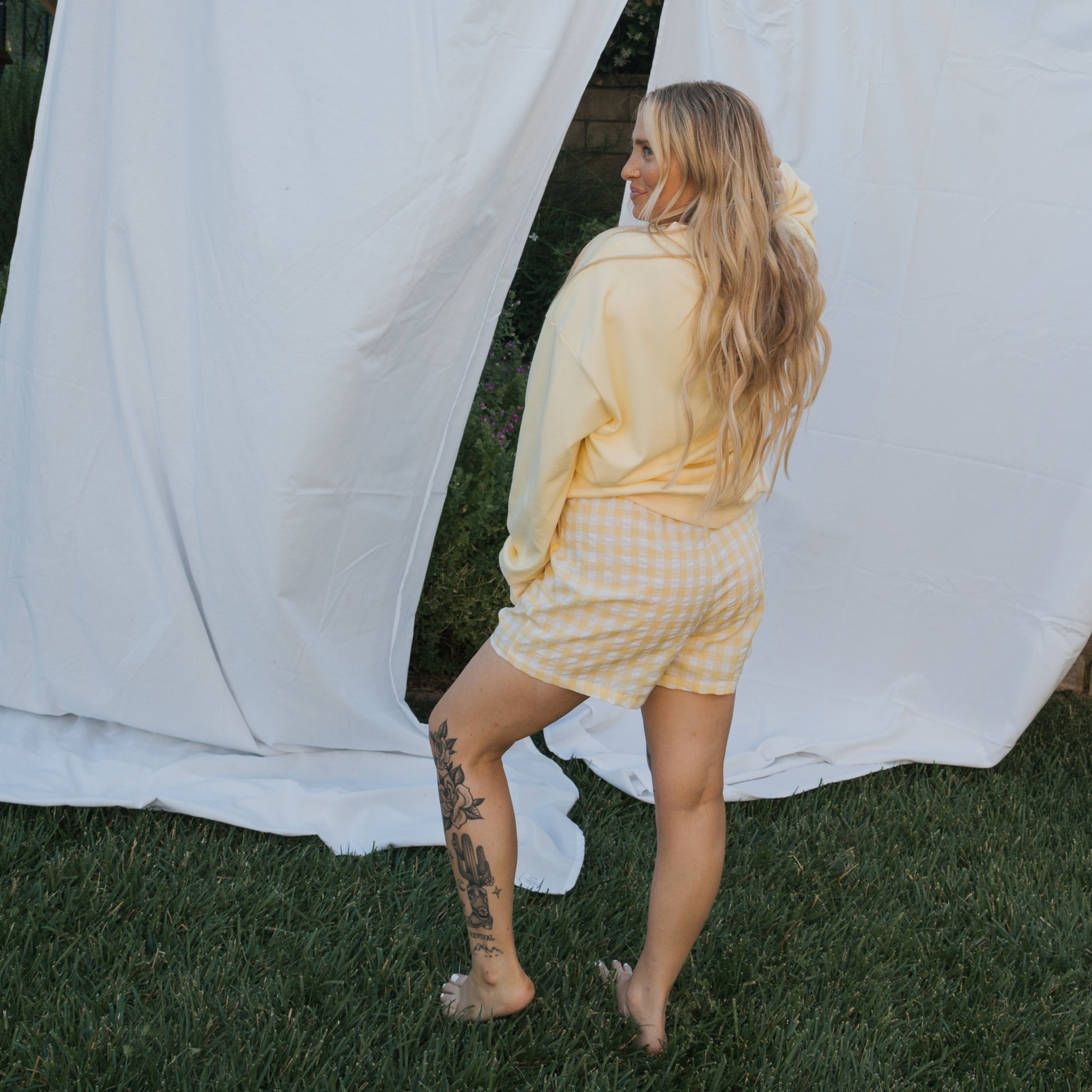 A woman with long blonde hair stands barefoot on grass, wearing a yellow sweatshirt and forever french baby Women's Boxer Shorts | Citron. She has a floral tattoo on her leg and gazes to the side by white fabric hanging outdoors.