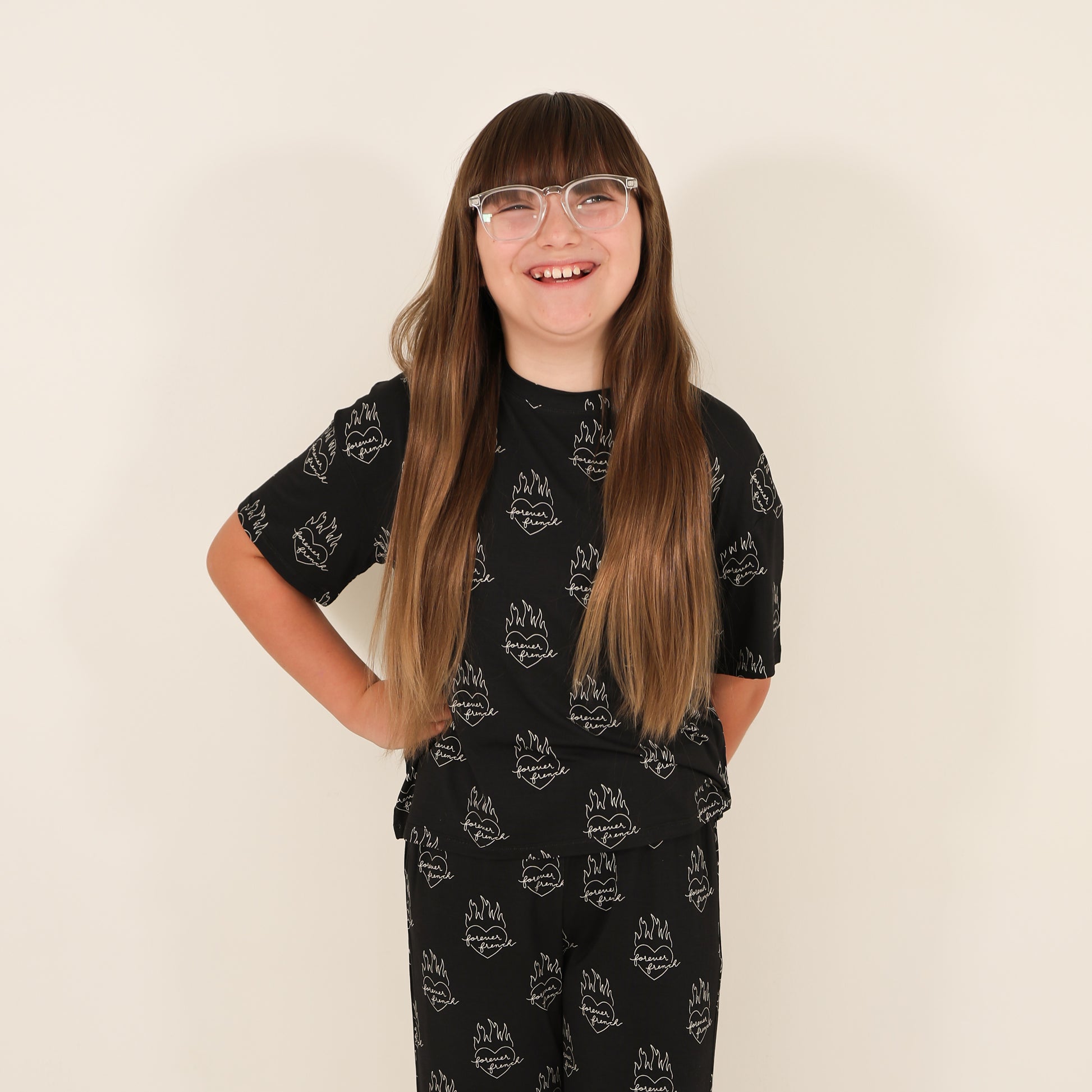 A smiling child with long brown hair and glasses wears the "Child Boxy Tee Lounge Set | Flaming Hearts" by forever french baby, featuring matching black bamboo pieces with a white flame design against a plain light background.