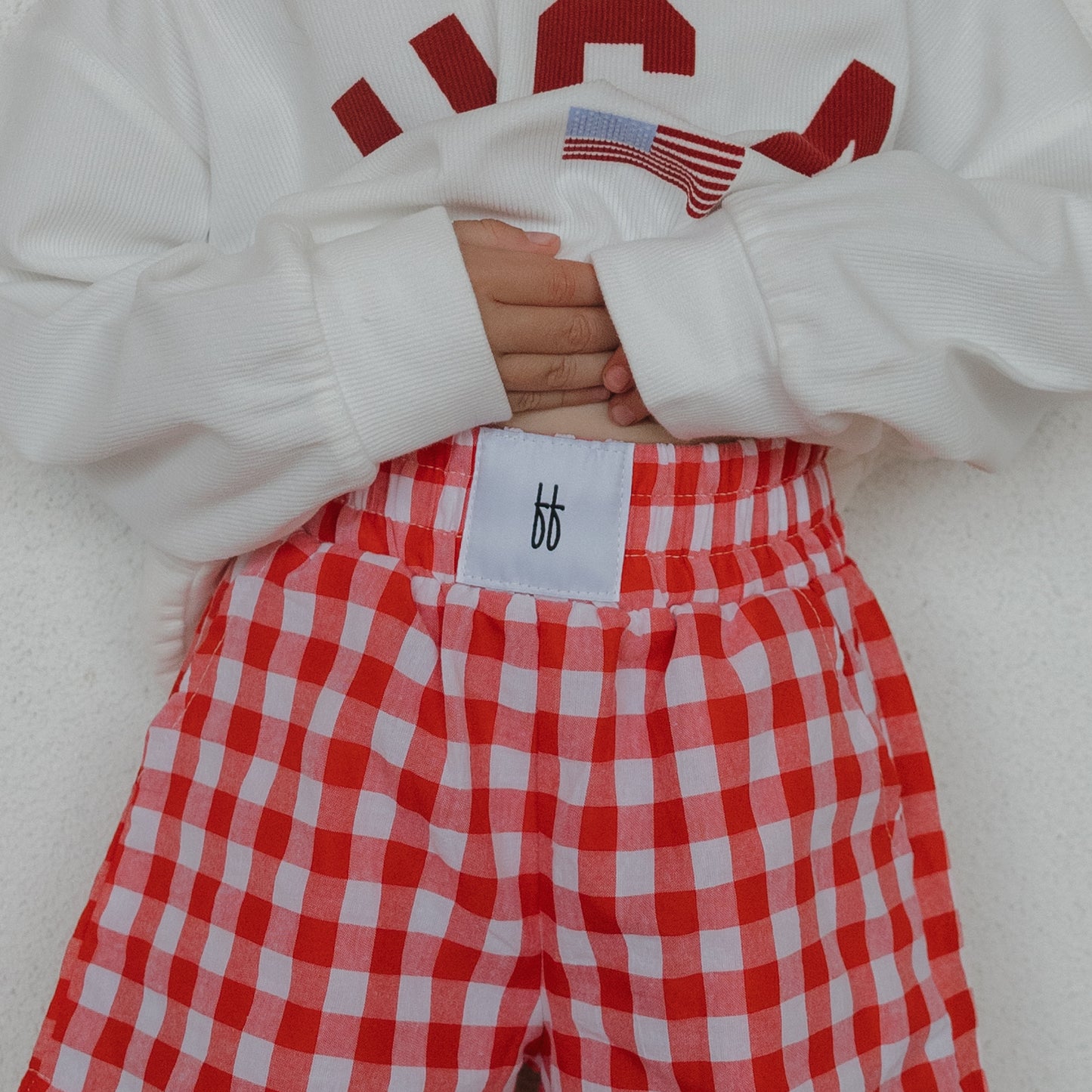 A child wears forever french baby’s Child Boxer Shorts in Scarlet Gingham, paired with a white 100% cotton sweatshirt featuring red lettering, standing against a light background with hands on their stomach.