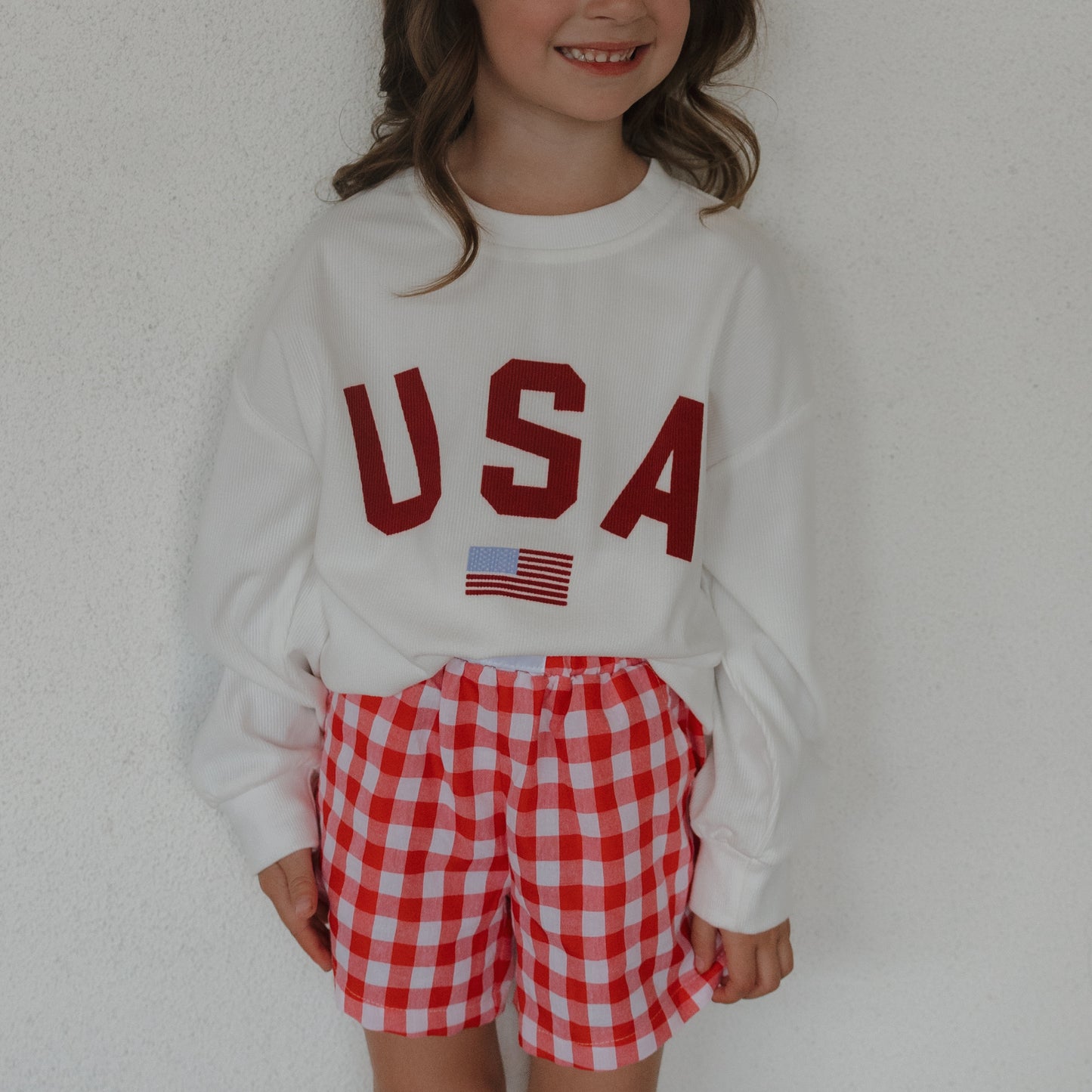 A young child smiles in a white "USA" sweatshirt and forever french baby Child Boxer Shorts | Scarlet Gingham, made from 100% cotton, standing against a plain white background.