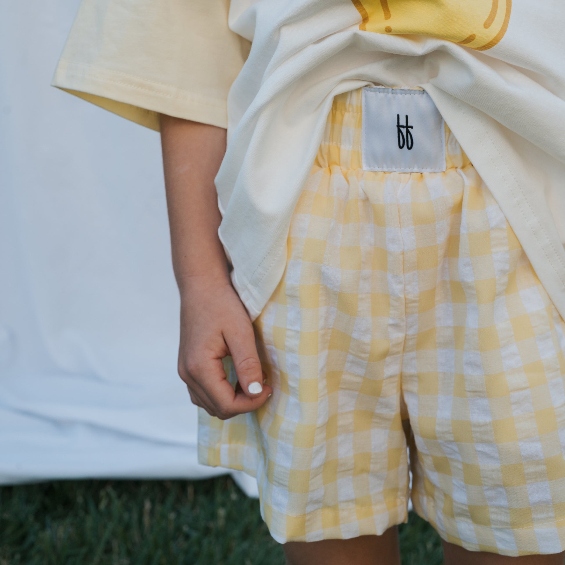 A child stands on grass in forever french baby Child Boxer Shorts | Citron, paired with a cream tee. Only their torso and part of their legs are visible, with one hand relaxed at their side.