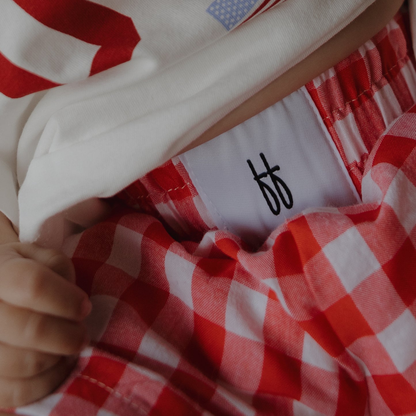A child's hand holds the waistband of forever french baby Child Boxer Shorts | Scarlet Gingham, showing a white tag with an "ff" logo. The 100% cotton shorts are paired with a white shirt decorated in red and blue.