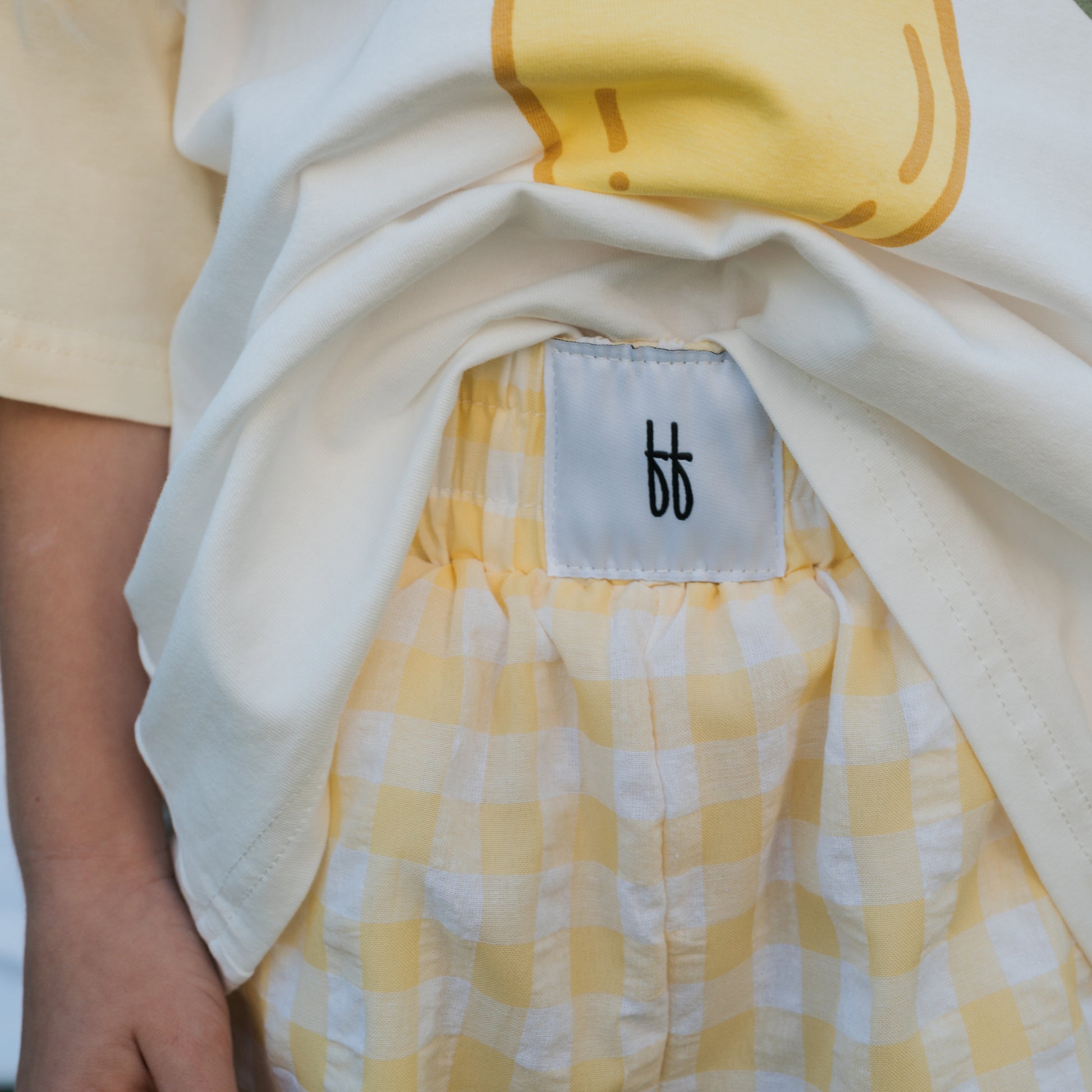 A child lifts a white shirt to reveal forever french baby Child Boxer Shorts | Citron—100% cotton, yellow and white checks, elastic waistband, and a white tag with the double “b” logo.