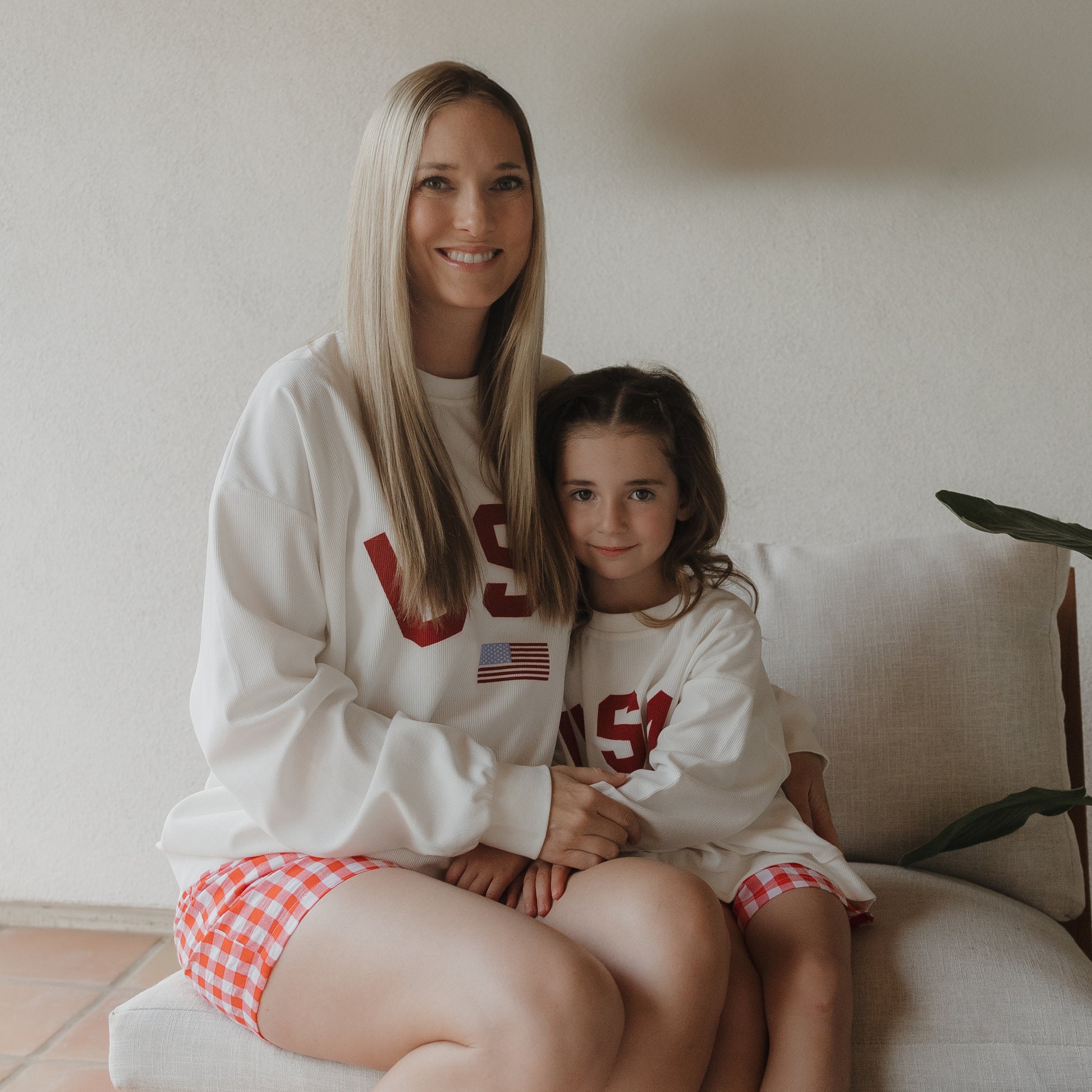 A woman and a young girl sit on a light chair, smiling and holding hands. Both wear matching white USA sweatshirts and forever french baby Women's Boxer Shorts in Scarlet Gingham. A plant sits beside them.
