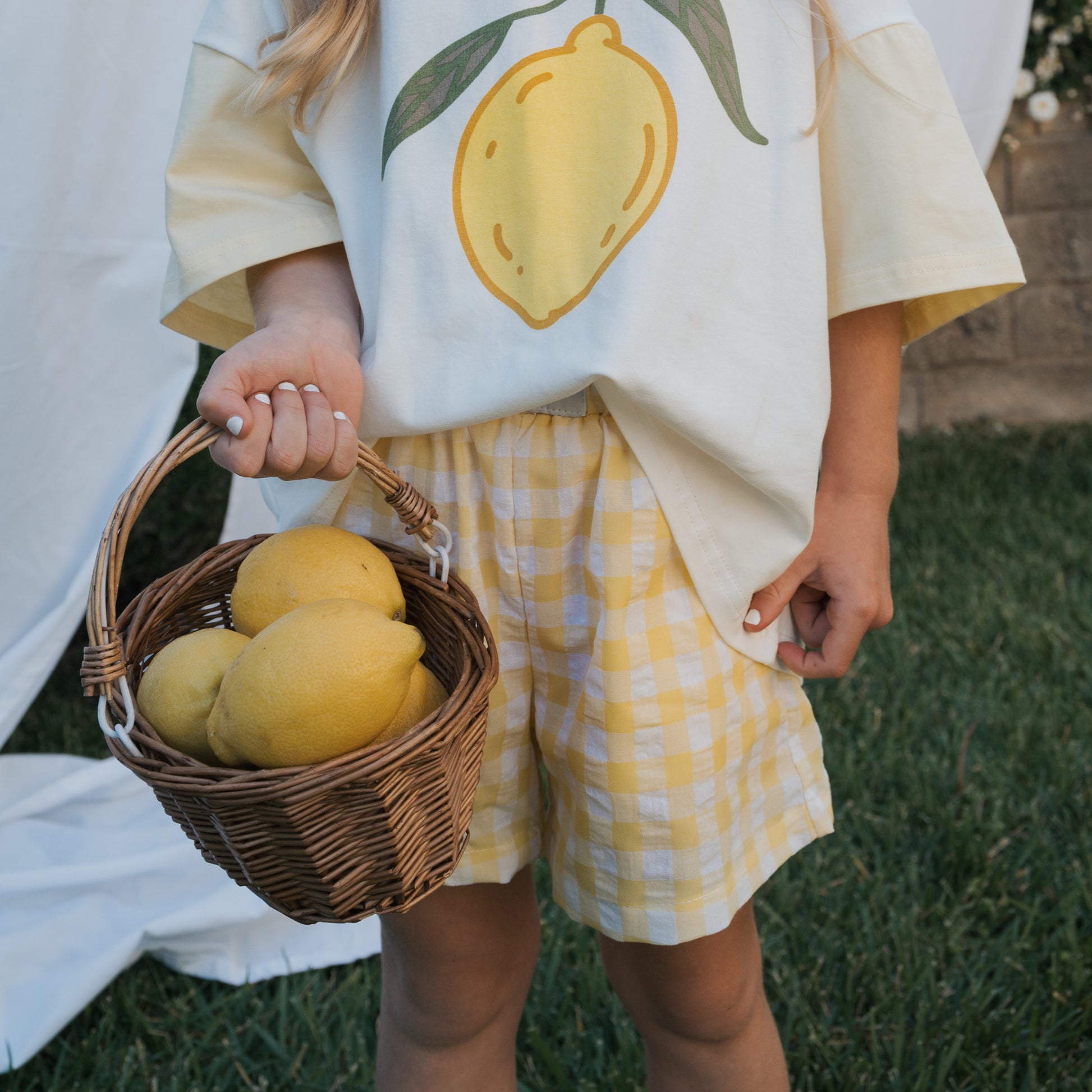 A child in forever french baby’s 100% cotton lemon-print shirt and Child Boxer Shorts | Citron stands on grass holding a basket of lemons.