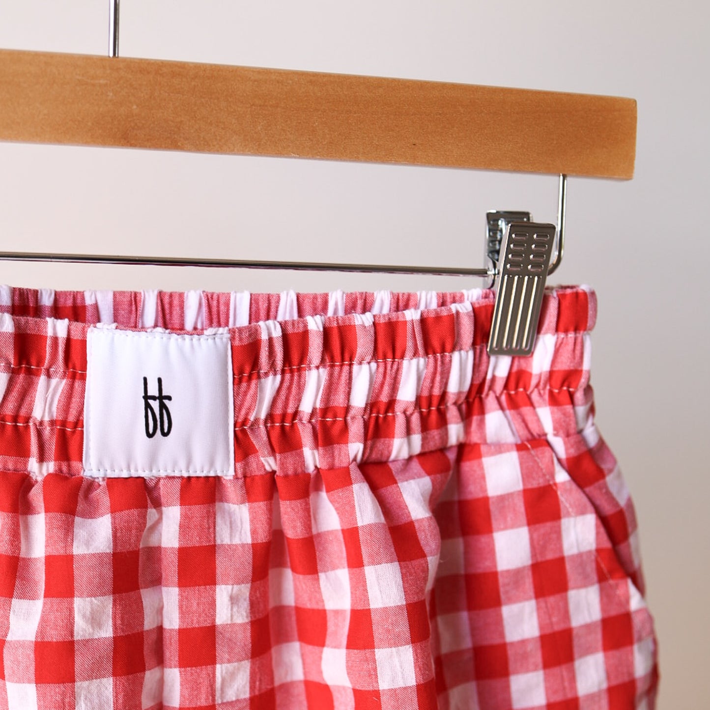 Women's Boxer Shorts | Scarlet Gingham by forever french baby, in red and white gingham 100% cotton with relaxed fit and elastic waistband, hang on a wooden hanger. A white label with black "bb" logo is visible; background is plain and neutral.