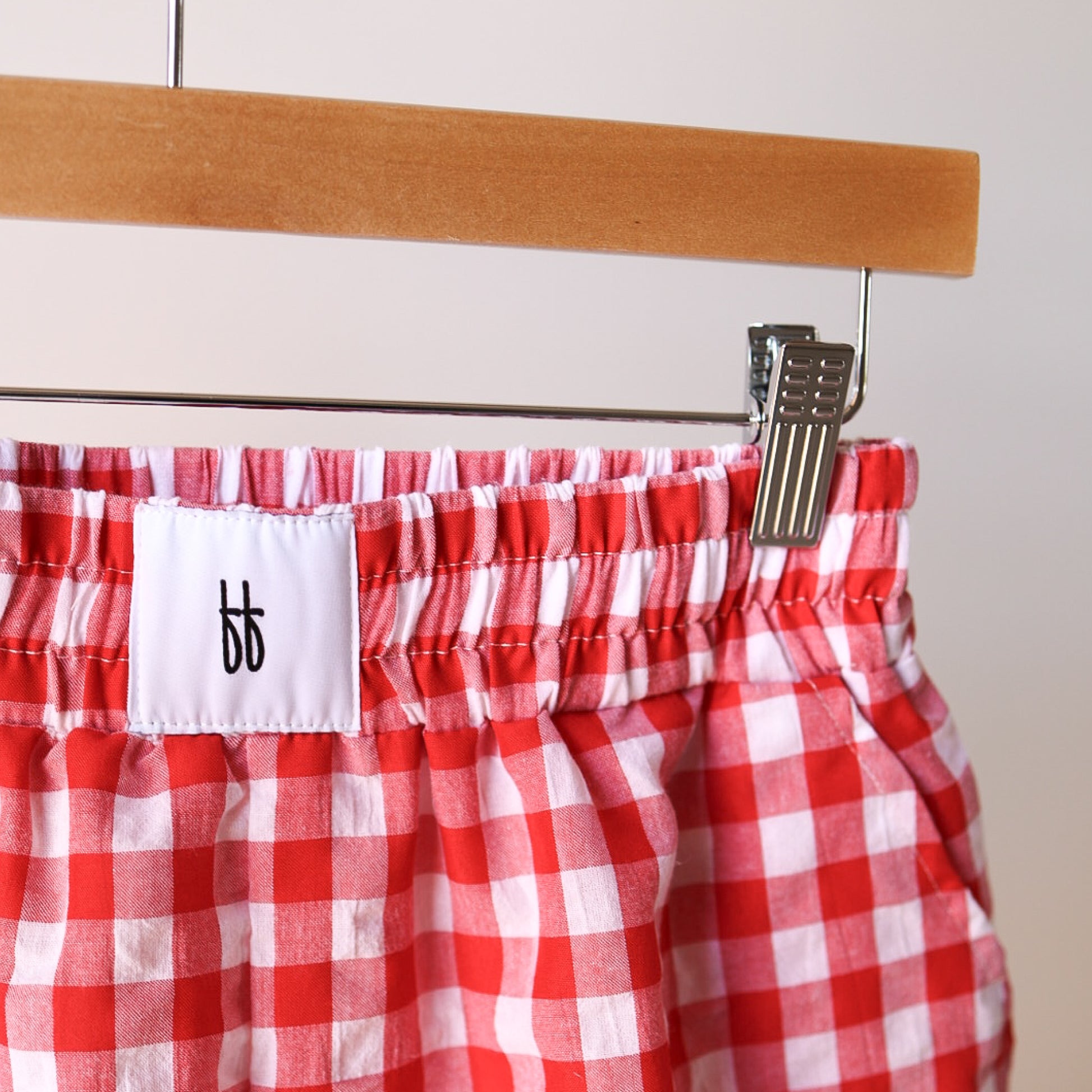 Women's Boxer Shorts | Scarlet Gingham by forever french baby, in red and white gingham 100% cotton with relaxed fit and elastic waistband, hang on a wooden hanger. A white label with black "bb" logo is visible; background is plain and neutral.