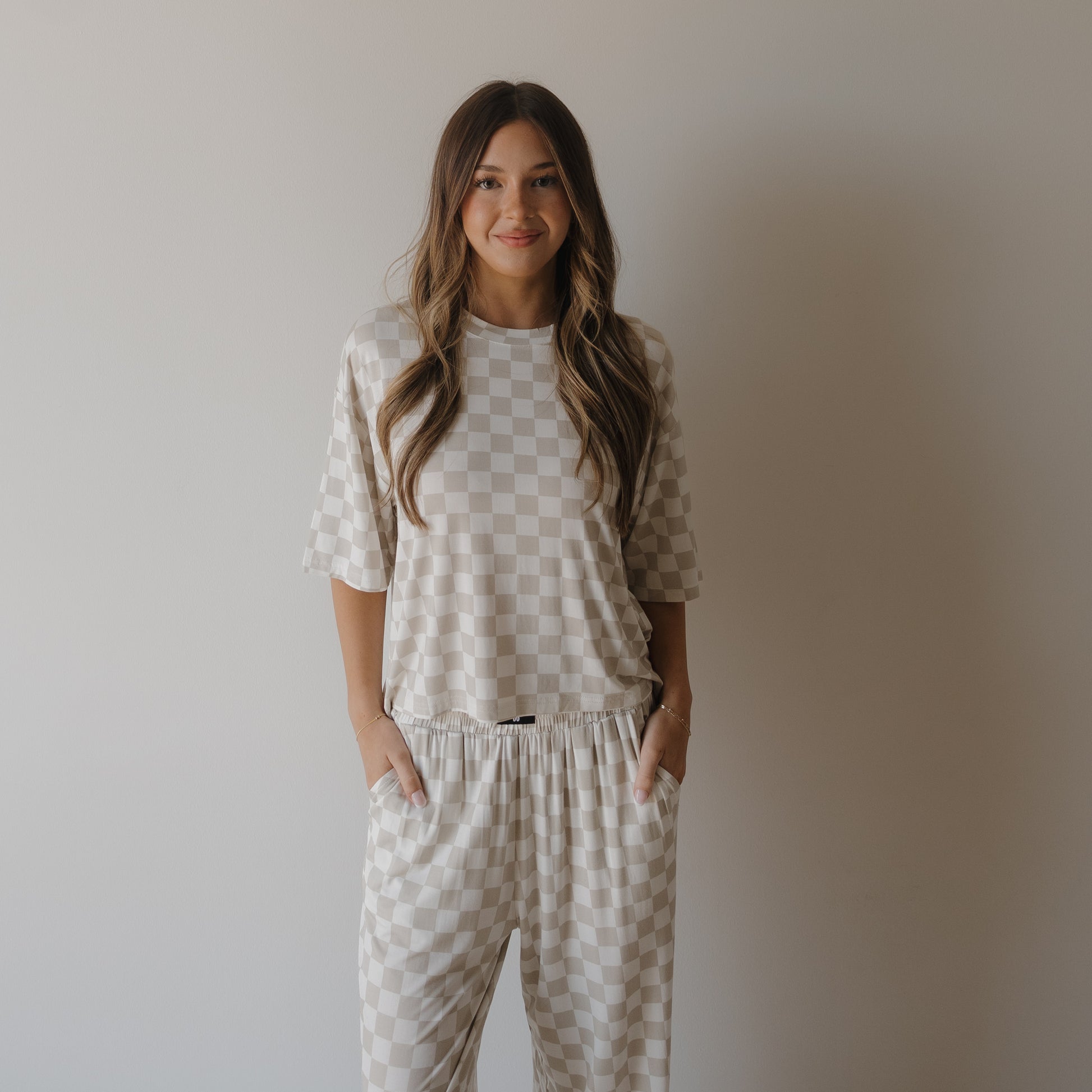 A woman with long wavy hair smiles with her hands in her pockets, modeling the Women's Boxy Tee Lounge Set | Checkerboard by forever french baby. She stands against a plain light background, highlighting the comfort of this bamboo clothing set.
