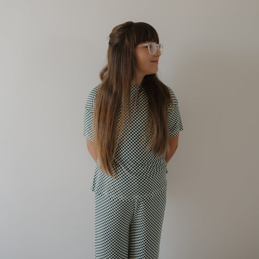 A young girl with long brown hair and glasses wears the forever french baby Child Boxy Tee Lounge Set in Lucky Micro Check, standing against a plain white wall, looking to her left with hands behind her back.