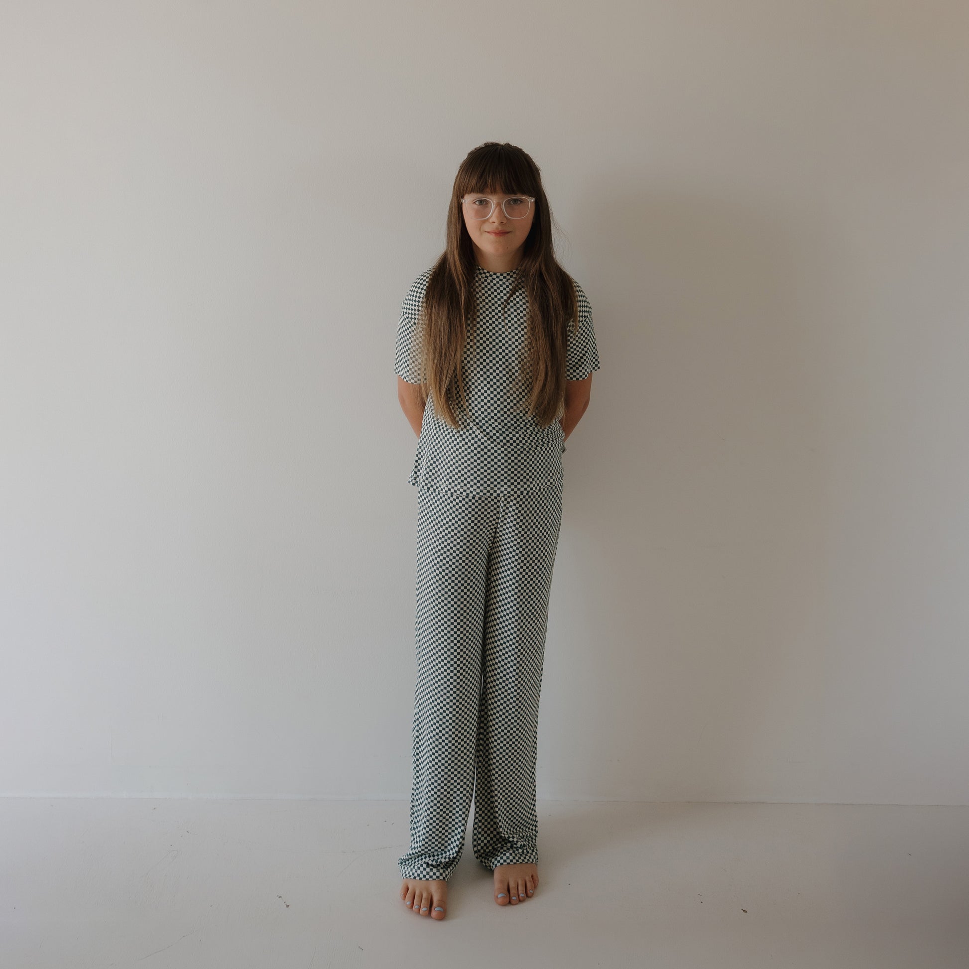 A young girl with long brown hair and glasses stands barefoot against a white wall, wearing the forever french baby Child Boxy Tee Lounge Set in Lucky Micro Check. Her hands are behind her back.