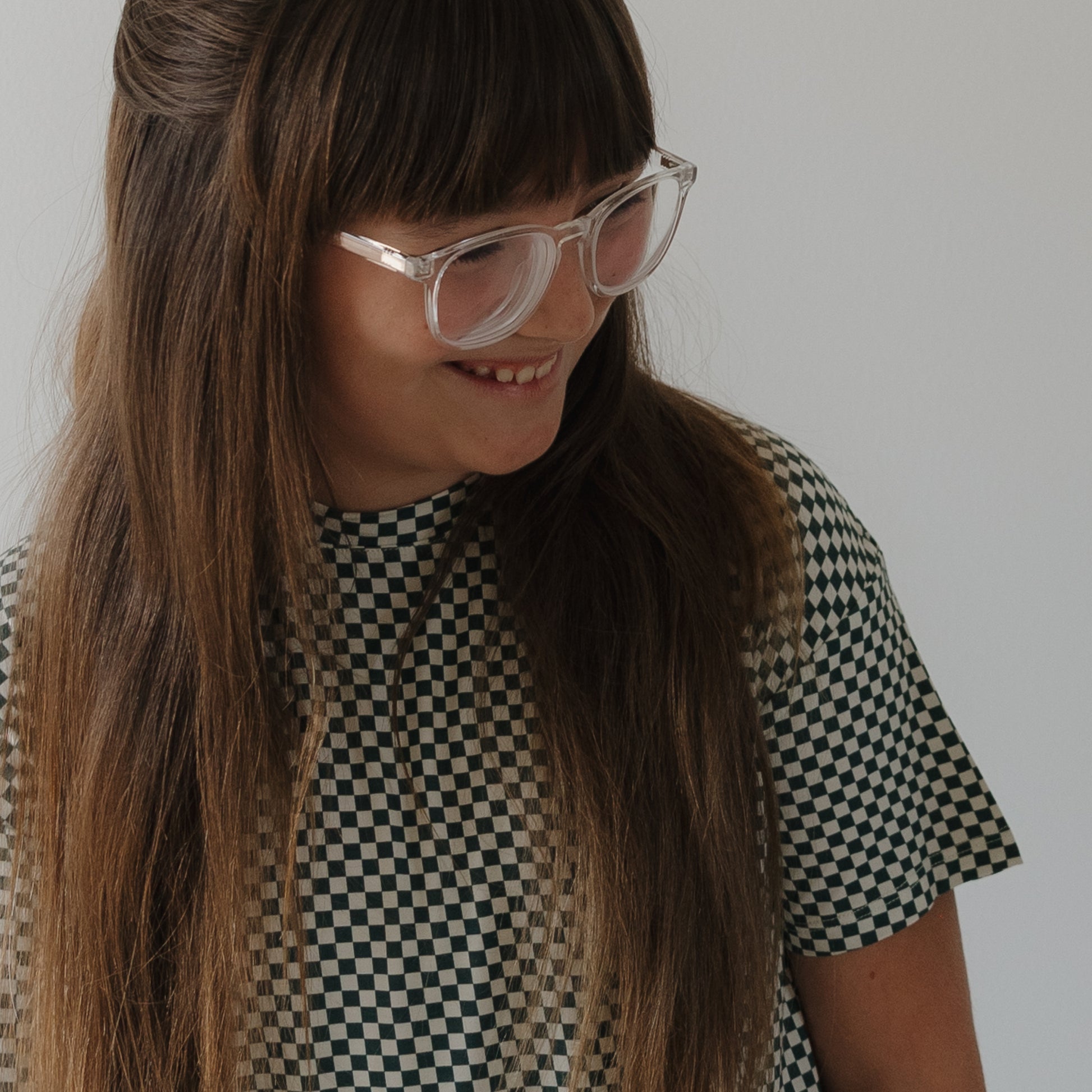 A child with long brown hair and bangs, wearing large clear glasses and the forever french baby Child Boxy Tee Lounge Set in Lucky Micro Check, smiles while looking down against a plain light background.