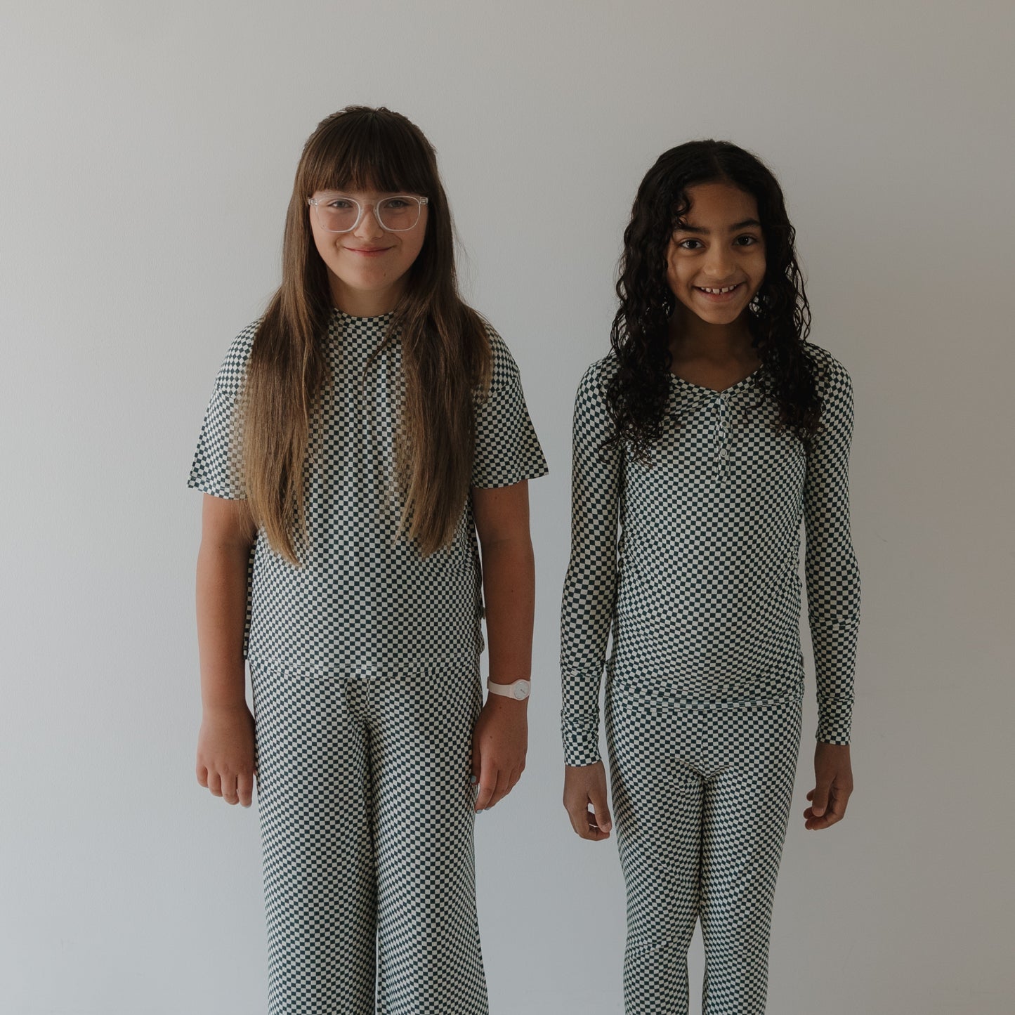 Two young girls smile side by side in forever french baby's Bamboo Two Piece Pajamas | Lucky Micro Check, made from hypo-allergenic fabric that's perfect for sensitive skin. One wears glasses with straight hair; the other has long curly hair.