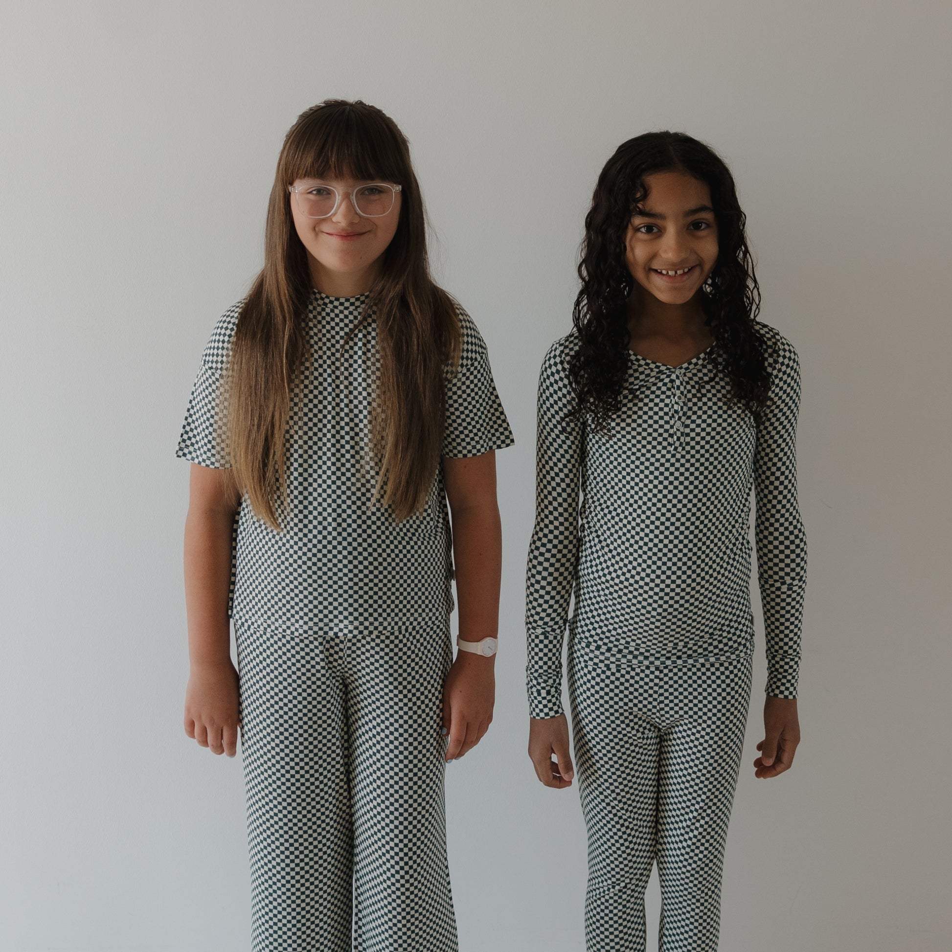 Two young girls smile side by side in forever french baby's Bamboo Two Piece Pajamas | Lucky Micro Check, made from hypo-allergenic fabric that's perfect for sensitive skin. One wears glasses with straight hair; the other has long curly hair.