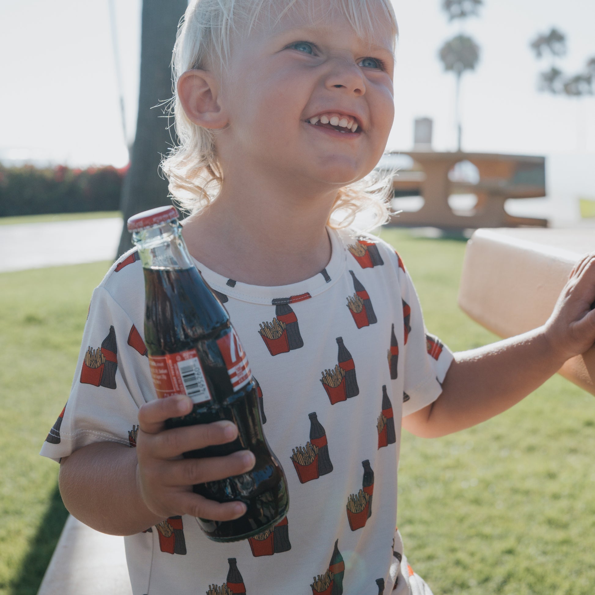 A smiling blond child in forever french baby's Bamboo Short Set | Fries & Cola sits on a bench in a sunny park, holding a glass bottle of Coca-Cola. Palm trees and a picnic table are seen behind the child.