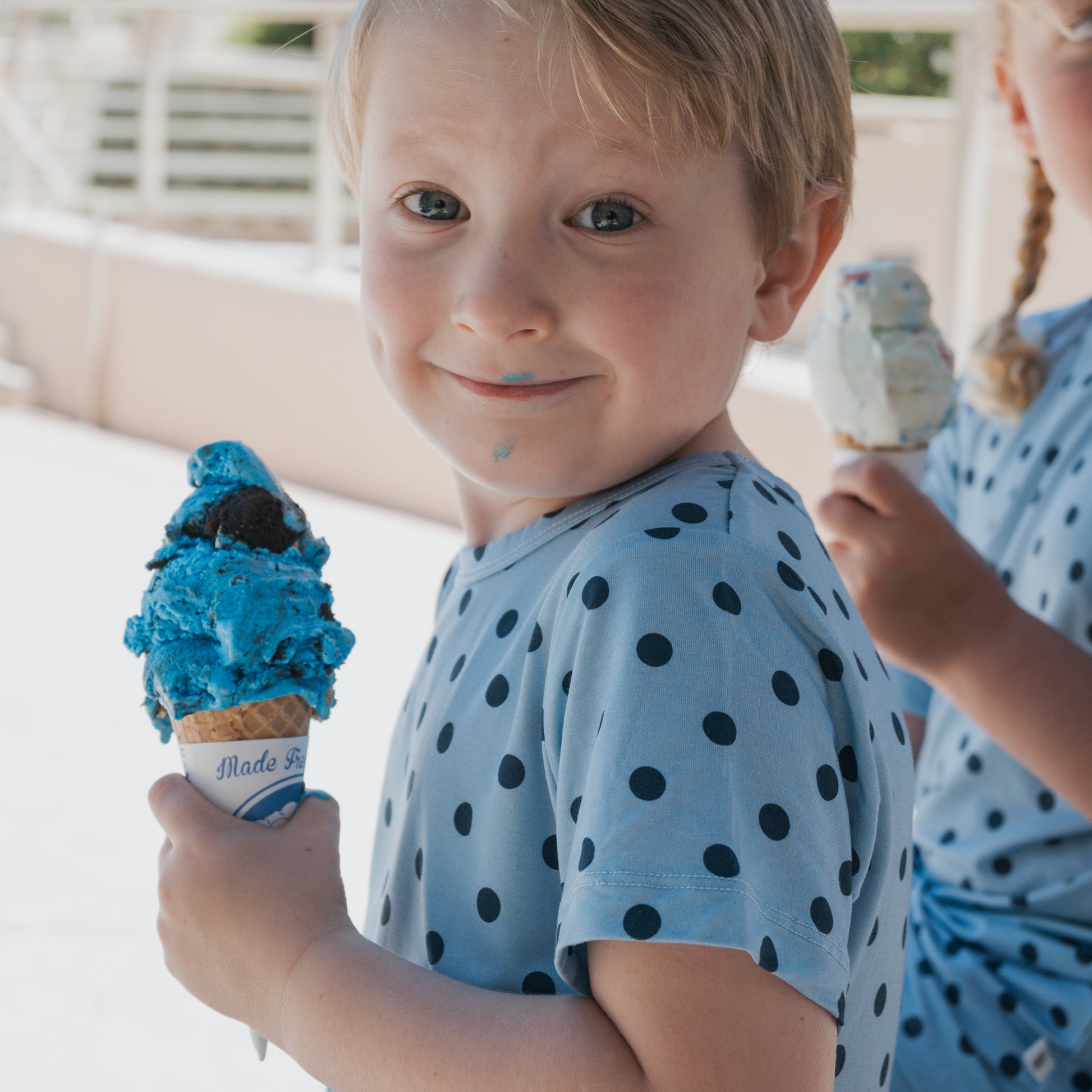 A smiling child with blonde hair, blue ice cream on their lips, wears the forever french baby Bamboo Short Set | Sully—stylish pajamas crafted from hypoallergenic bamboo, gentle for sensitive skin. Their light blue shirt features dark polka dots. Another child holding an ice cream cone appears partially in the background.