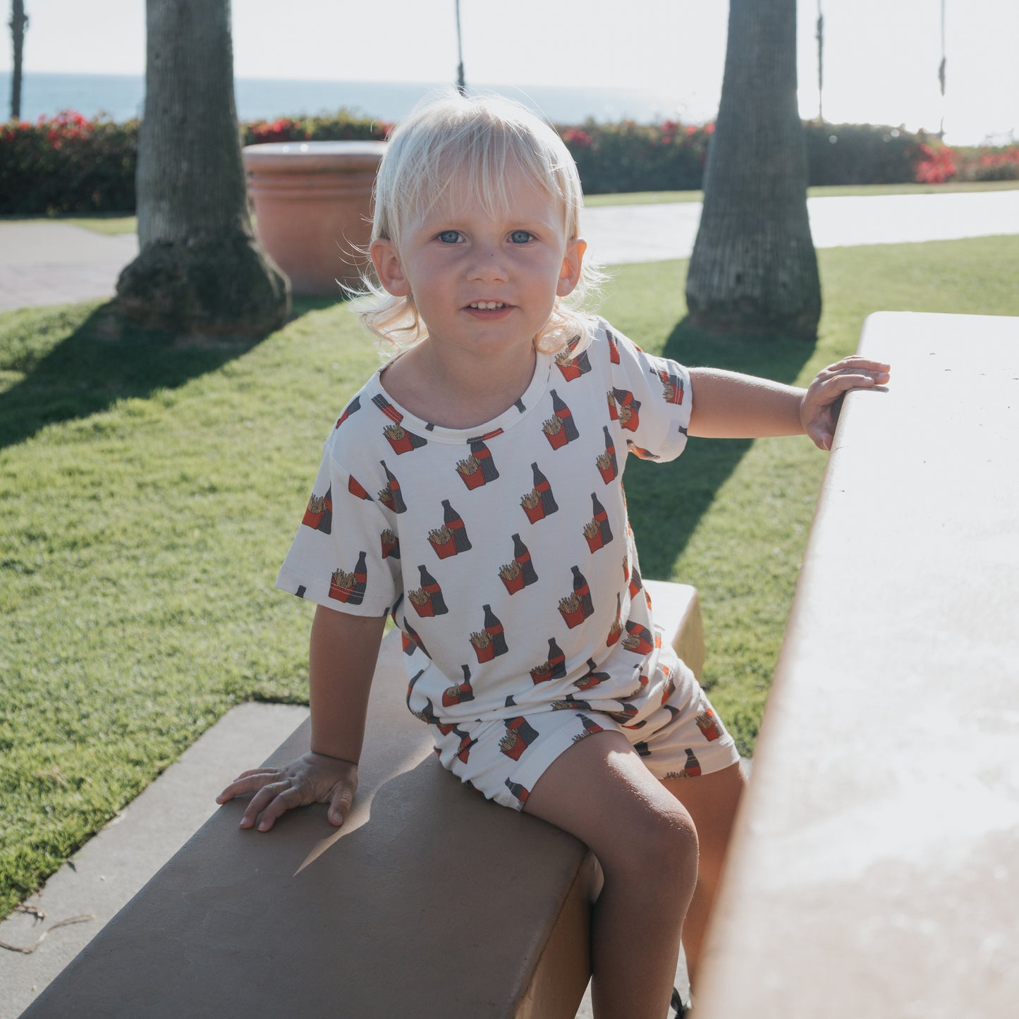 A young child with blonde hair sits on a bench outdoors, wearing the forever french baby Bamboo Short Set | Fries & Cola. Bright sunlight, green grass, and palm trees are visible in the background.