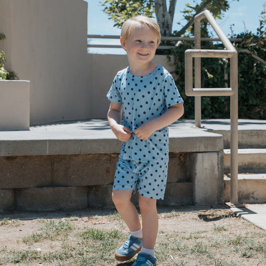 A young child wearing the forever french baby Bamboo Short Set in the Sully print smiles while standing outside on a sunny day near concrete steps, grass, and a metal railing.
