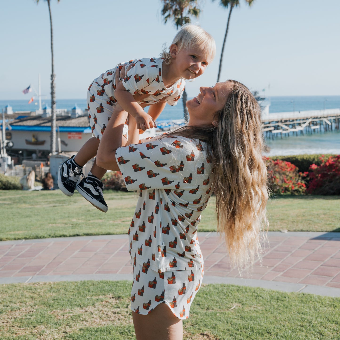 A woman with long blonde hair lifts a smiling toddler in the air. Both wear matching forever french baby Bamboo Short Sets | Fries & Cola, featuring a brown pattern, outdoors by grass and the ocean on a sunny day.