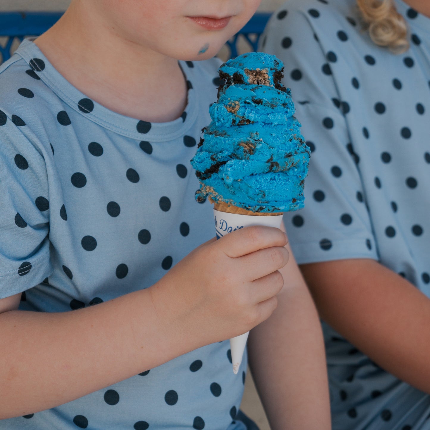 A child wears the Bamboo Short Set | Sully by forever french baby—a blue polka dot shirt made from breathable bamboo—while holding a large blue ice cream cone. Beside them, another person in a matching set sits partially visible.