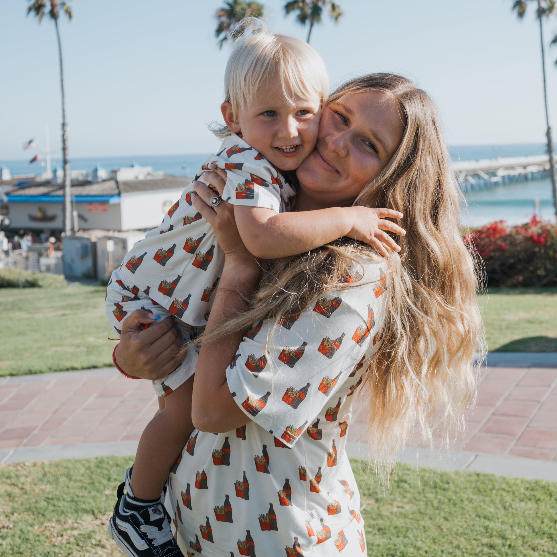 A smiling woman with long blonde hair holds a young child outdoors near the beach. Both wear forever french baby Bamboo Short Set | Fries & Cola—matching kids bamboo clothing perfect for sensitive skin. Palm trees and ocean are in the background.