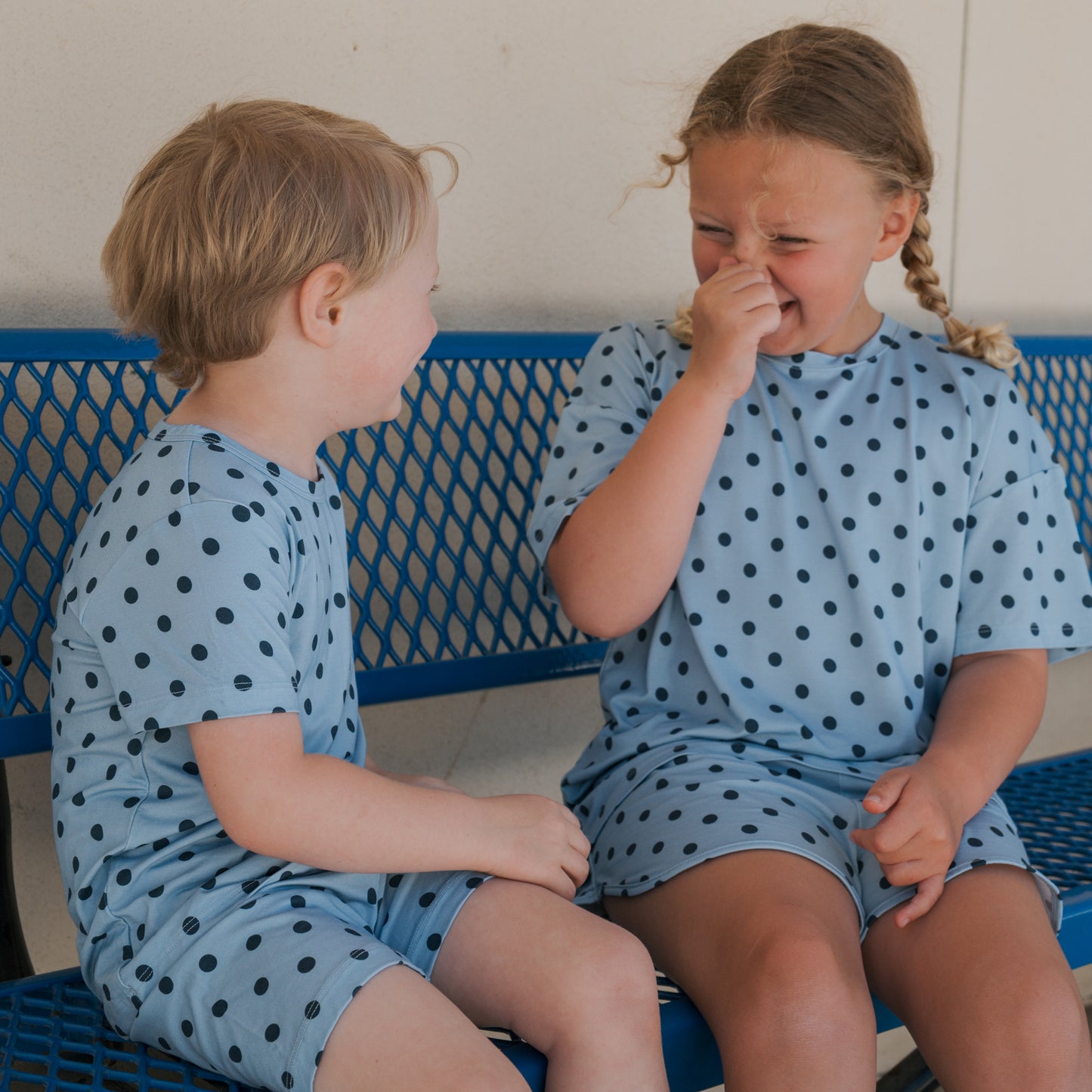 Two children sit on a blue bench outdoors against a light-colored wall, both dressed in matching "Bamboo Short Set | Sully" outfits from forever french baby. Made from breathable bamboo sleepwear material, one child laughs and covers their nose while looking at the other, who is smiling.