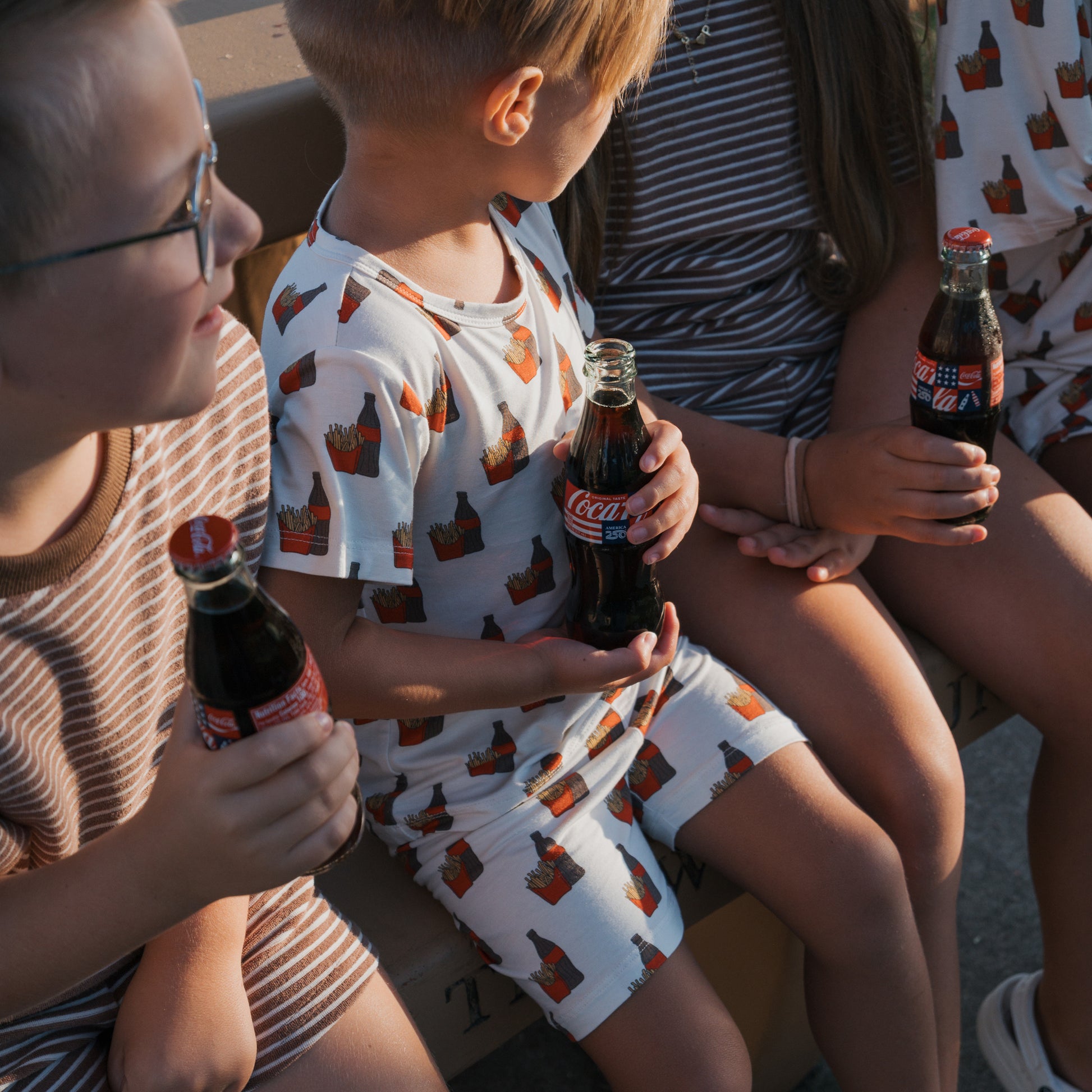 Four children in forever french baby’s Bamboo Short Set | Fries & Cola sit together, each holding a glass Coca-Cola bottle, dressed in soft, striped or patterned summery bamboo clothing as warm sunlight casts gentle shadows over them.