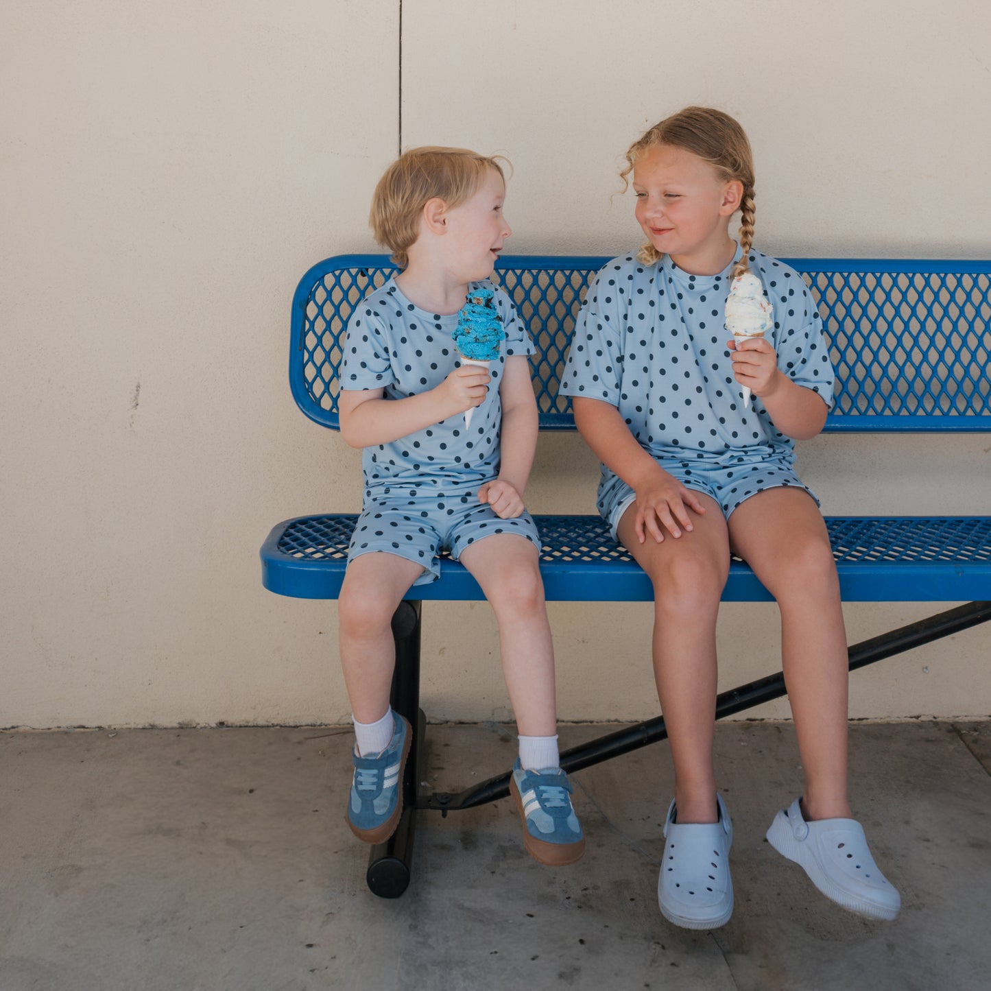 Two young children in forever french baby Bamboo Short Set | Sully pajamas sit on a blue metal bench, smiling at each other while holding ice cream cones.