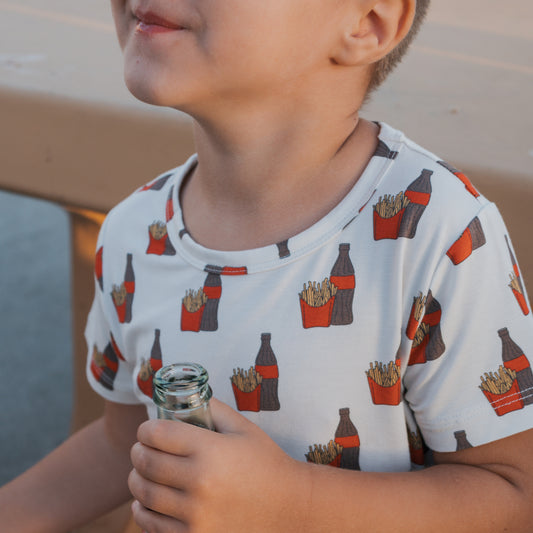 A young child in the forever french baby Bamboo Short Set | Fries & Cola sits at a tan table holding a glass soda bottle, wearing cozy clothing decorated with soda bottles and french fries. The child's face is partially visible.