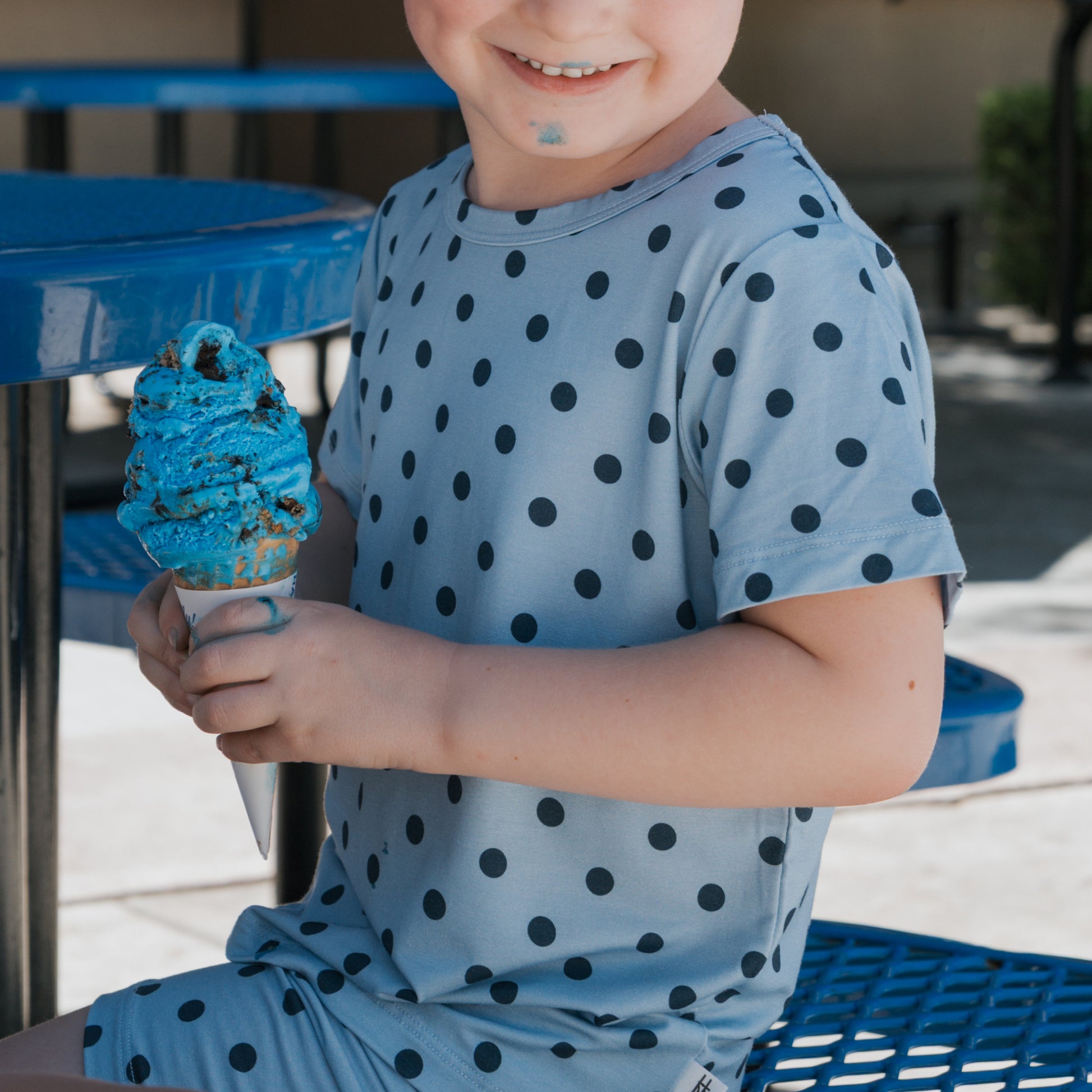 A smiling child, dressed in the forever french baby Bamboo Short Set | Sully, reminiscent of cozy hypoallergenic kids pajamas, holds a large blue ice cream cone with some ice cream on their chin while sitting at a blue outdoor table.