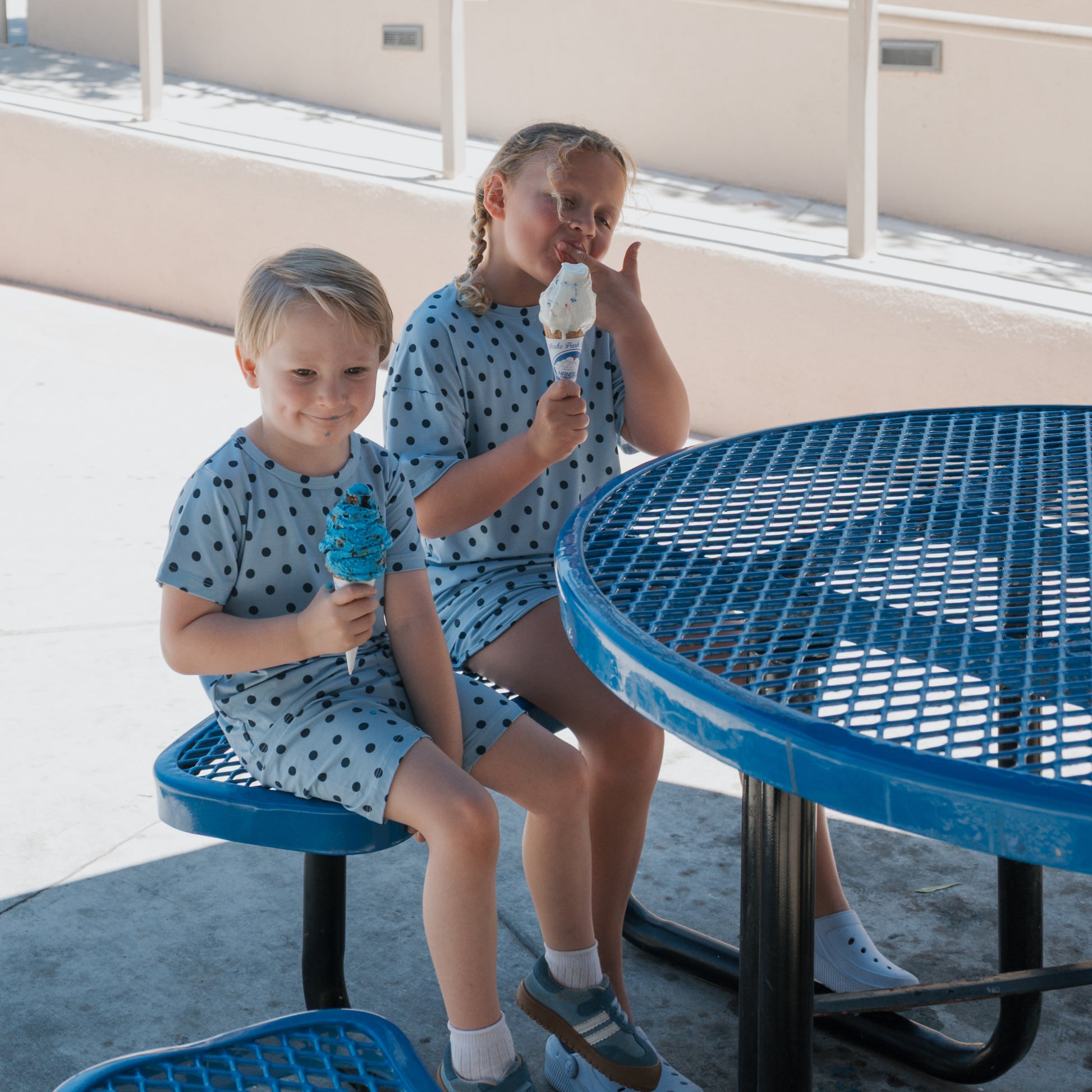 Two children sit at a blue metal picnic table on a sunny day, each holding an ice cream cone. The girl, wearing the Girl's Bamboo Short Set | Sully from forever french baby, is licking her ice cream while the boy smiles at the camera.