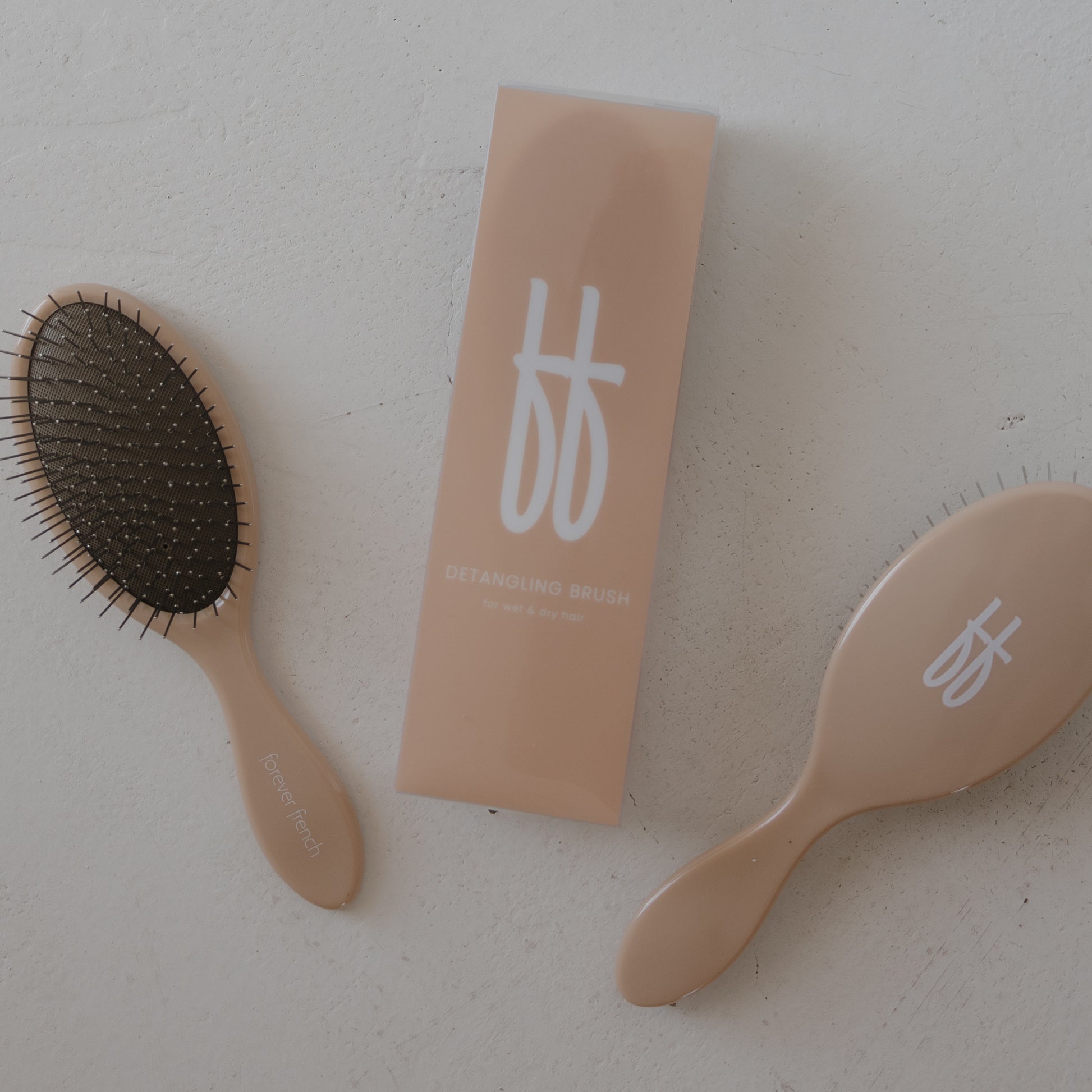 Two beige ff Hair Brushes by forever french baby, each with stainless steel bristles and a white stylized logo, are placed beside a matching rectangular box labeled "ff Hair Brush" on a light surface. The box and brushes feature the same logo.