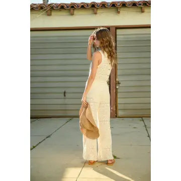 In front of a closed garage door, a woman with long hair stands wearing the MIOU MUSE Crochet Pattern Pants, a sleeveless white cotton jumpsuit featuring intricate crochet details. Holding a wide-brimmed hat in her left hand, she seems to be gazing into the distance.