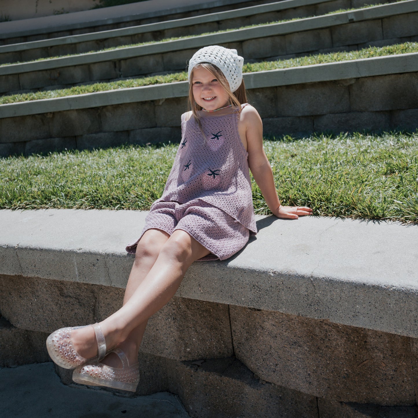 A young girl sits on a concrete ledge outdoors, smiling with her legs crossed. She wears a pink dress and the forever french baby Knit Head Scarf | Cloud, featuring an adjustable tie. Grass and stone steps are visible in the background.