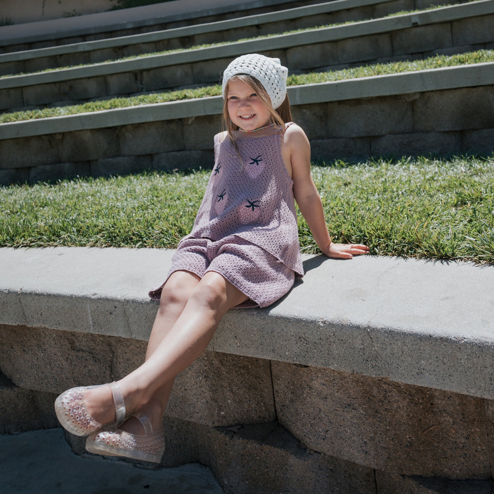 A young girl sits on a concrete ledge outdoors, smiling with her legs crossed. She wears a pink dress and the forever french baby Knit Head Scarf | Cloud, featuring an adjustable tie. Grass and stone steps are visible in the background.