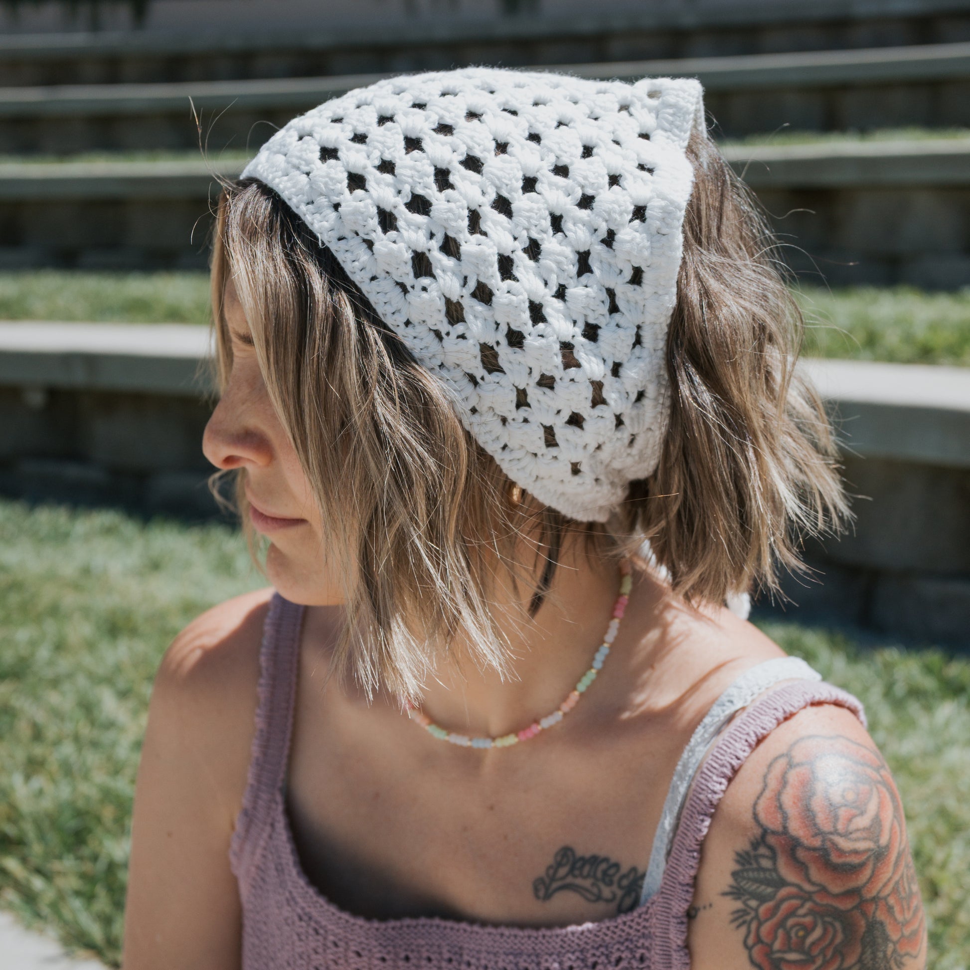 A woman with short, wavy hair is sitting outdoors on a sunny day with stone steps and grass in the background, wearing the forever french baby Knit Head Scarf | Cloud—a white cotton yarn crochet headscarf with an adjustable tie—along with a pink crocheted top, beaded necklace, and showing a visible arm tattoo.