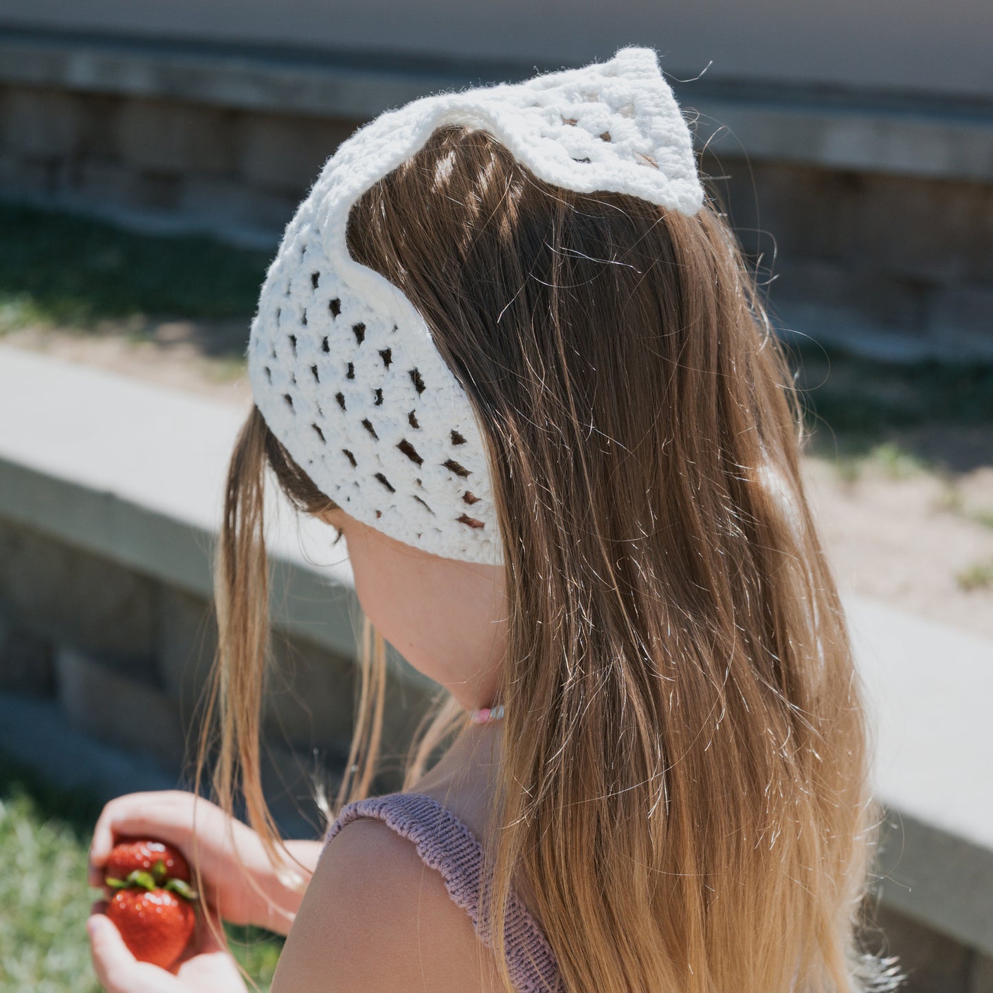A young girl with long hair wears the Knit Head Scarf | Cloud by forever french baby—crafted from cotton yarn with an adjustable tie securing it in place—as she holds strawberries outdoors on a sunny day.