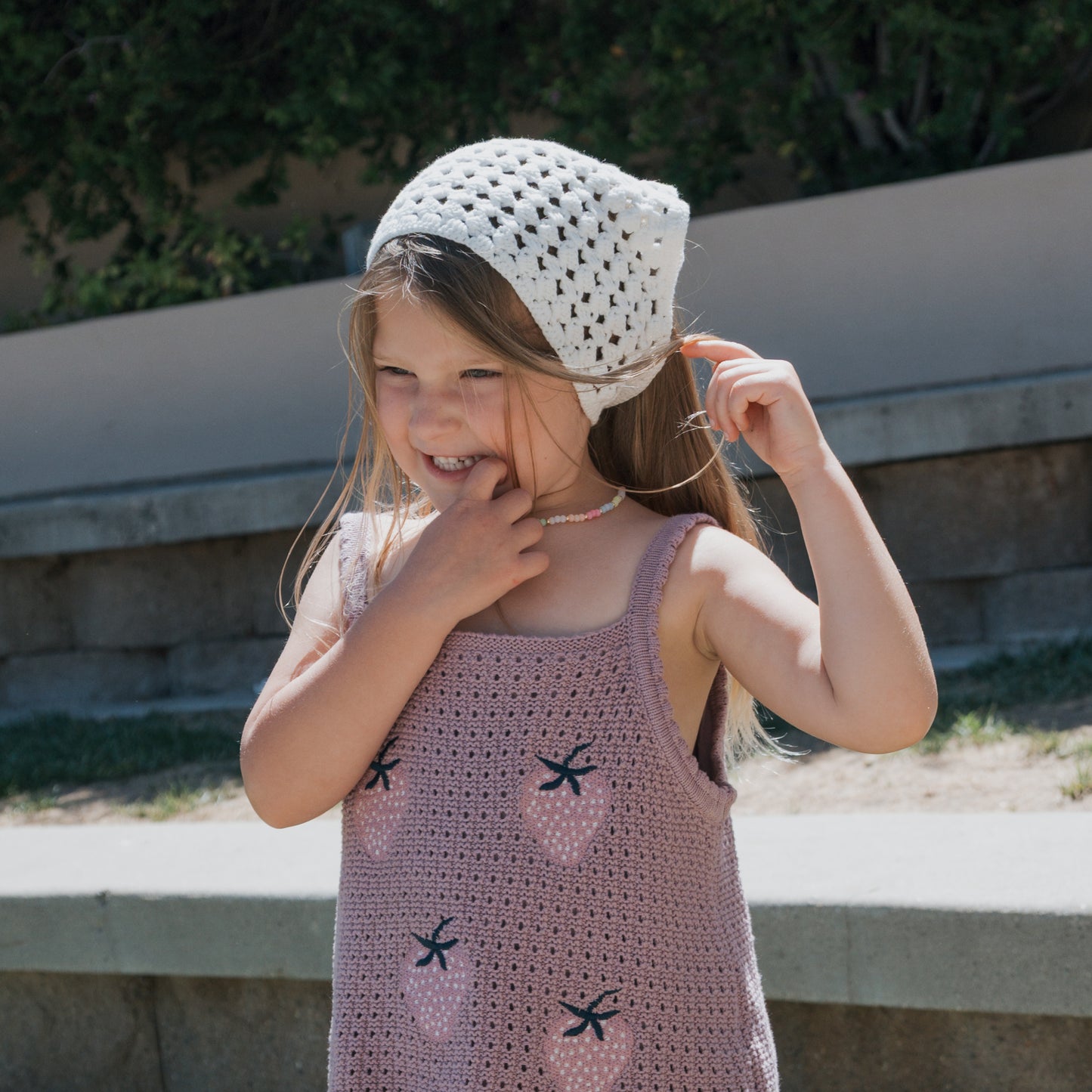 A young girl smiles outdoors, wearing a lavender crocheted dress decorated with strawberries, made from soft cotton yarn, and the Knit Head Scarf | Cloud by forever french baby—featuring an adjustable tie—surrounded by lush greenery in the background.