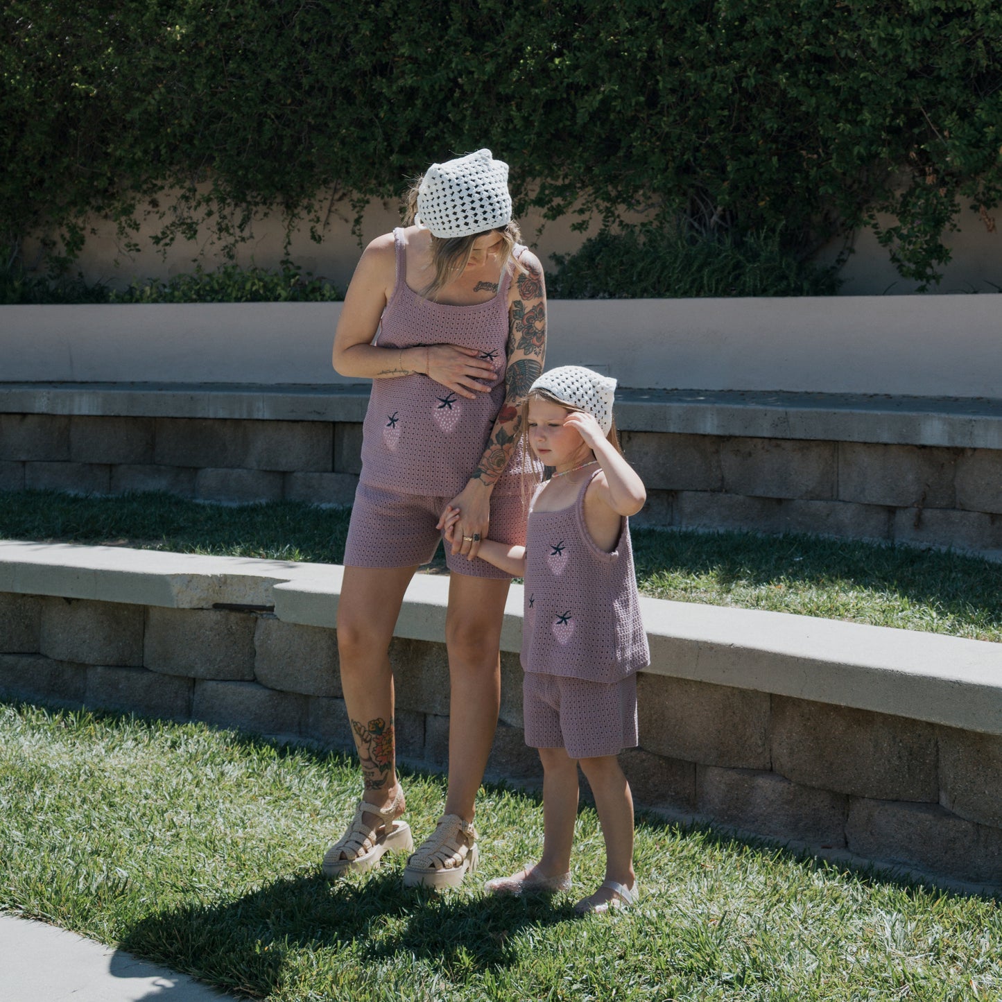 An adult and a child wearing matching purple cotton outfits and white "Knit Head Scarf | Cloud" headscarves from forever french baby stand on grass near stone seating, holding hands on a sunny day.