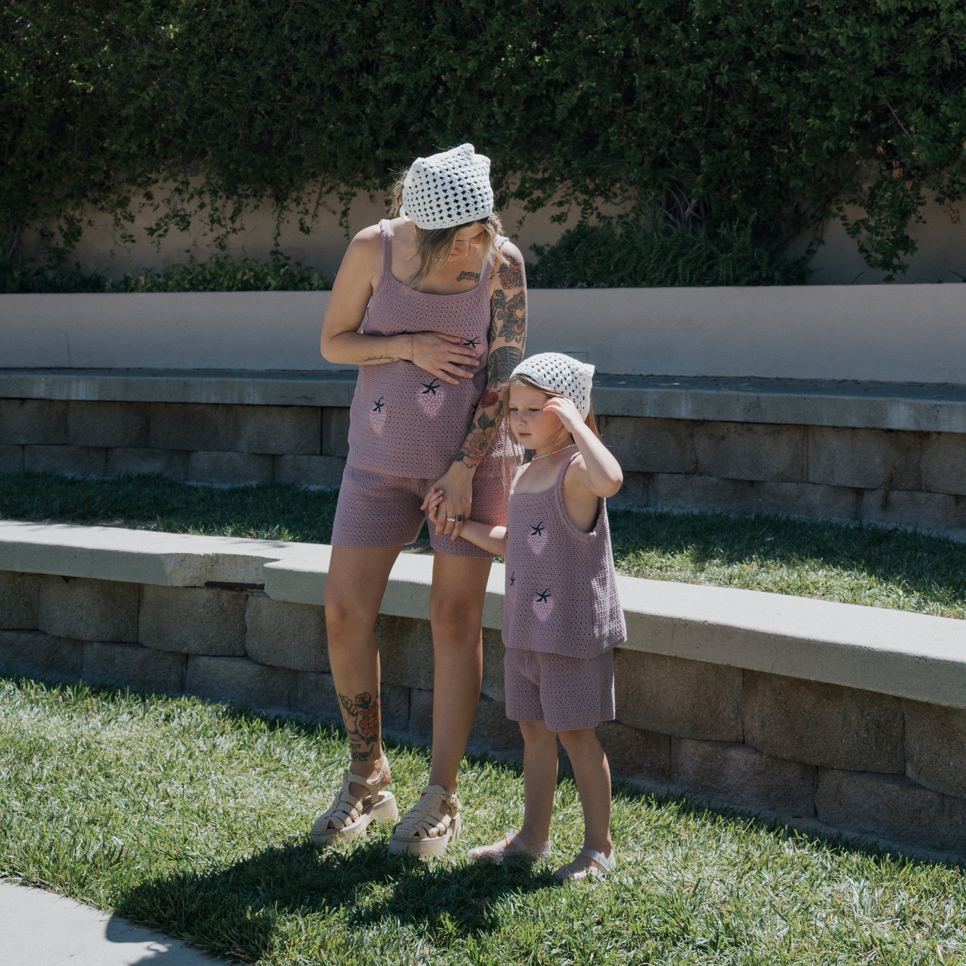 An adult and a child wearing matching purple cotton outfits and white "Knit Head Scarf | Cloud" headscarves from forever french baby stand on grass near stone seating, holding hands on a sunny day.