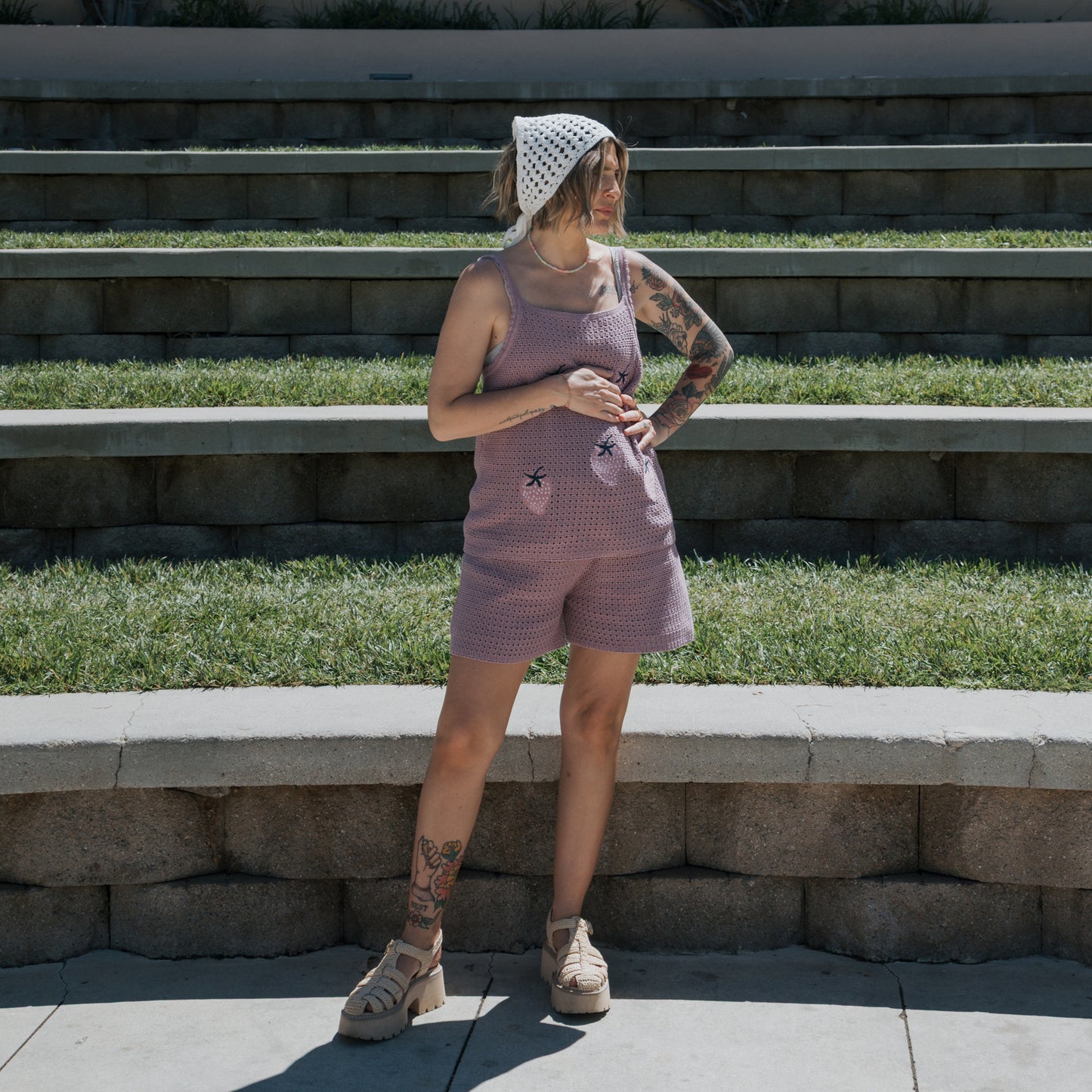 A person wearing the Knit Head Scarf | Cloud by forever french baby, a lavender crochet tank top crafted from soft cotton yarn, and shorts with purple sandals stands on a stone step outdoors, showcasing tattooed arms and legs. Grass and additional steps appear in the background.