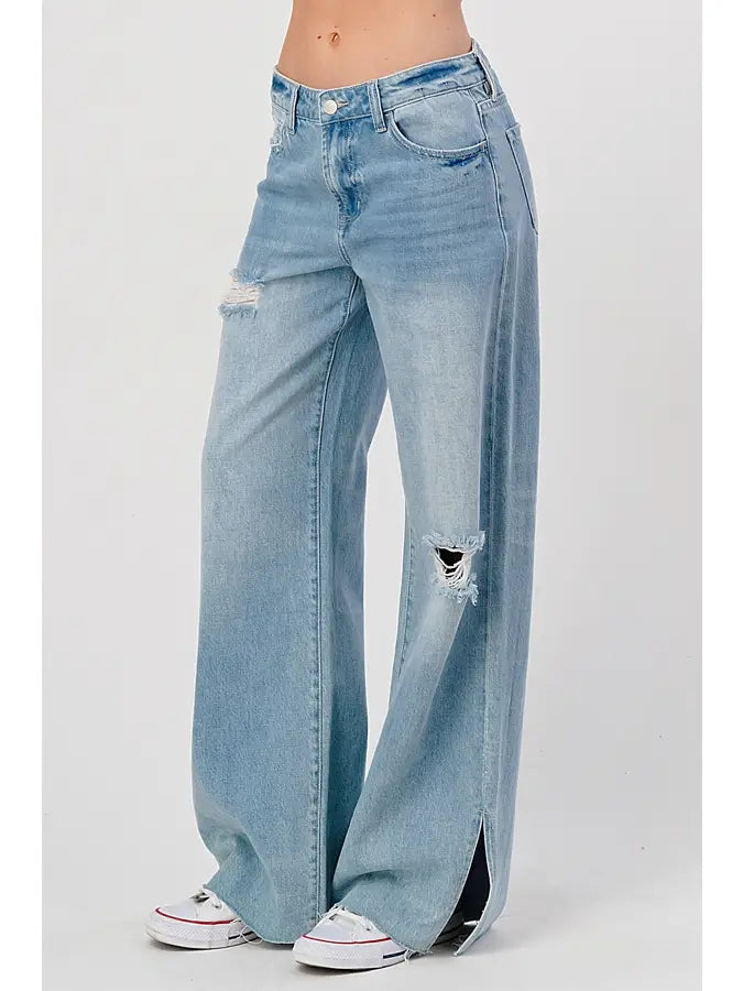 A person is wearing I&M JEAN, INC.'s High Rise Skater Jeans in medium wash with distressed detailing and wide leg openings, featuring small slits at the bottom. They are also sporting white sneakers with red accents.